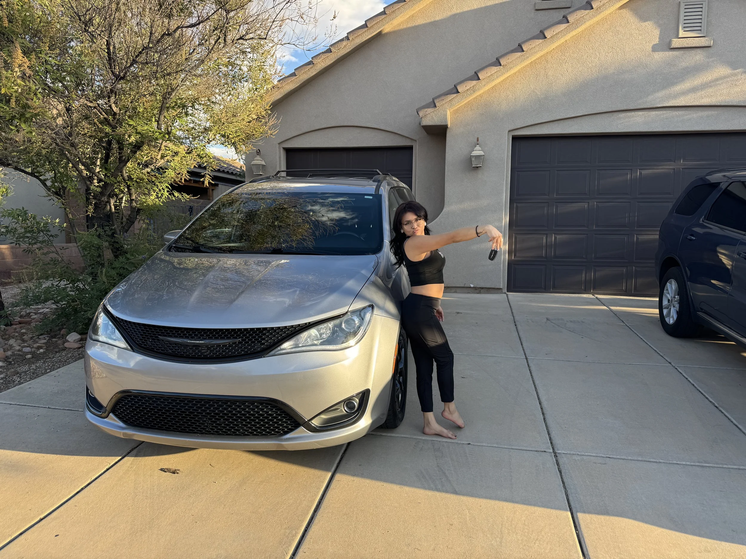 A woman in black clothing posing barefoot in front of a gray car (possibly a minivan) parked in a driveway, holding car keys in an extended arm. There is a house with a dark garage door and trees in the background.