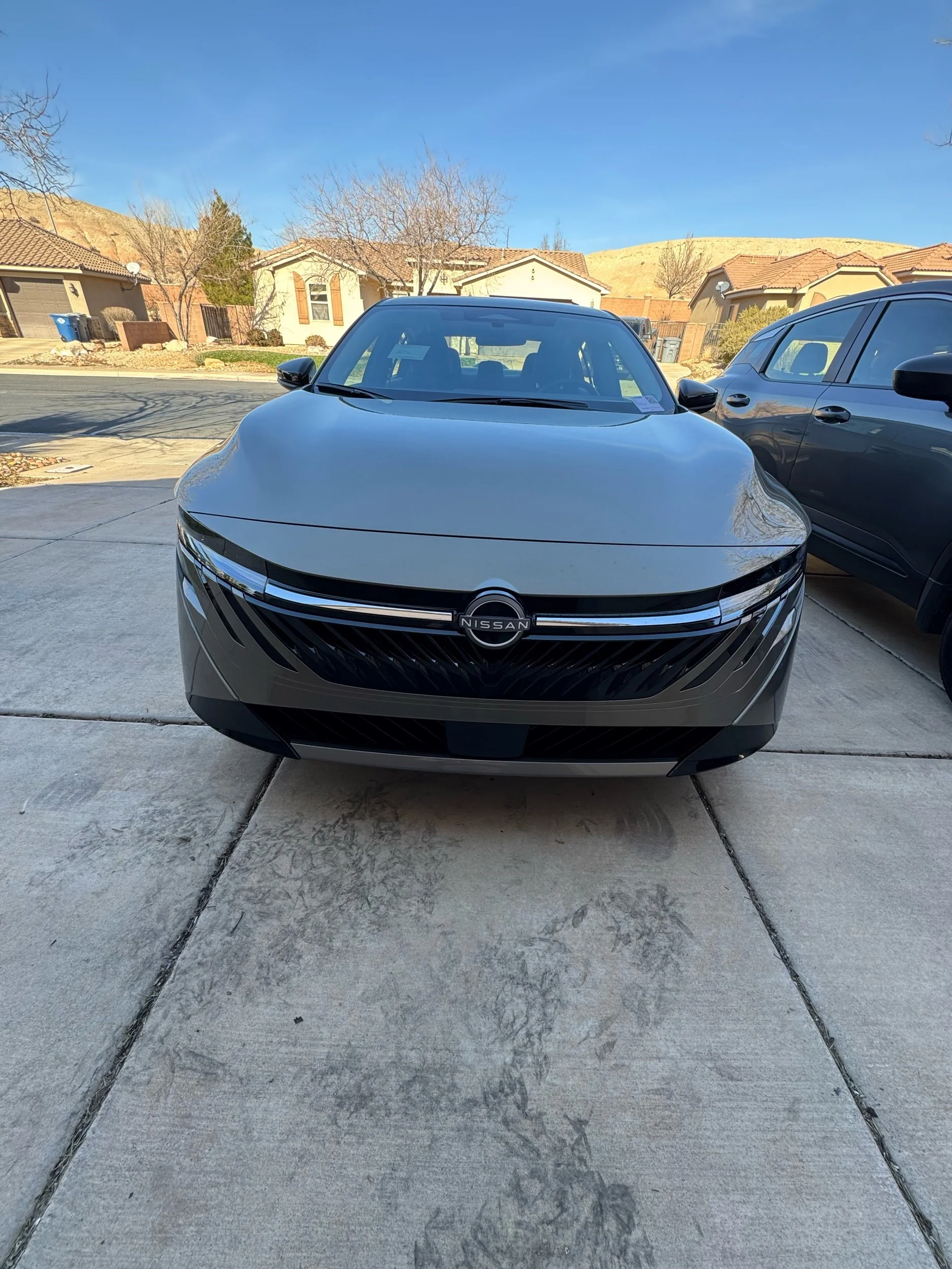 Front view of a black Nissan car parked on a concrete driveway in a residential neighborhood, with houses, leafless trees, and a clear blue sky in the background.