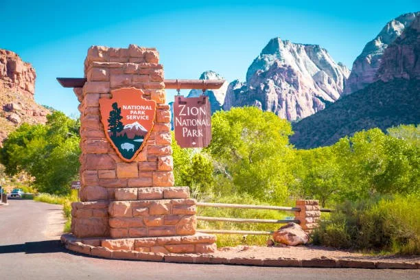 Entrance sign for Zion National Park with the park's logo and name, surrounded by green trees and mountains in the background.