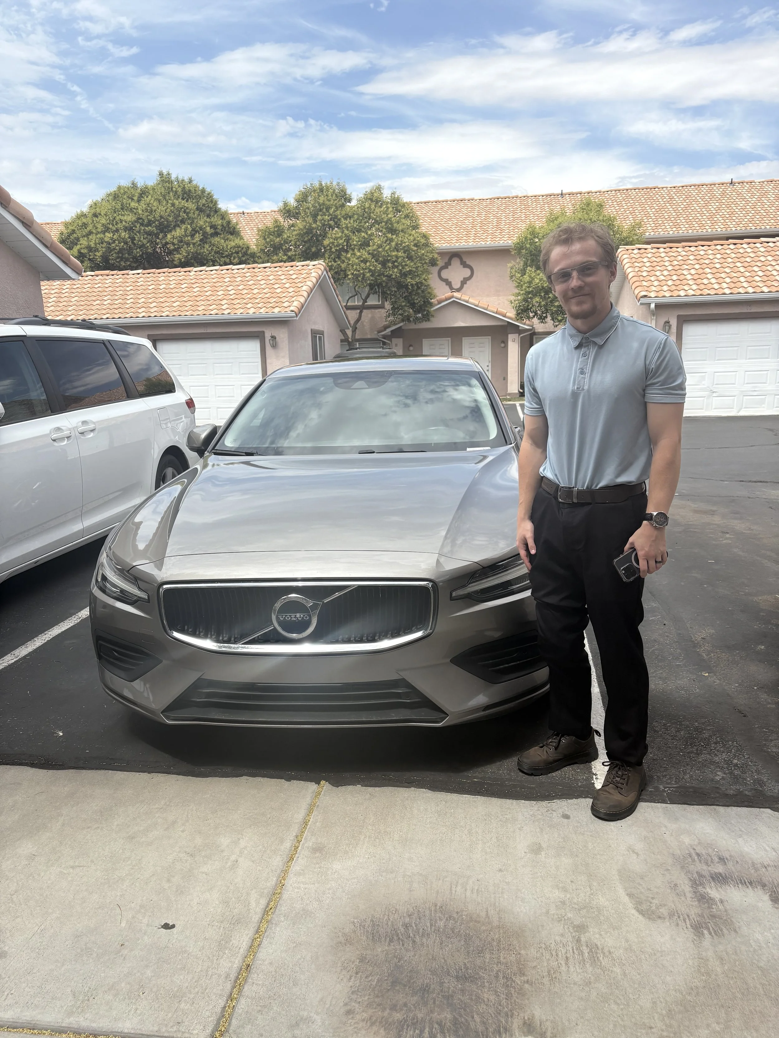 A man standing next to a silver Volvo sedan parked in a residential area with beige houses and trees in the background.