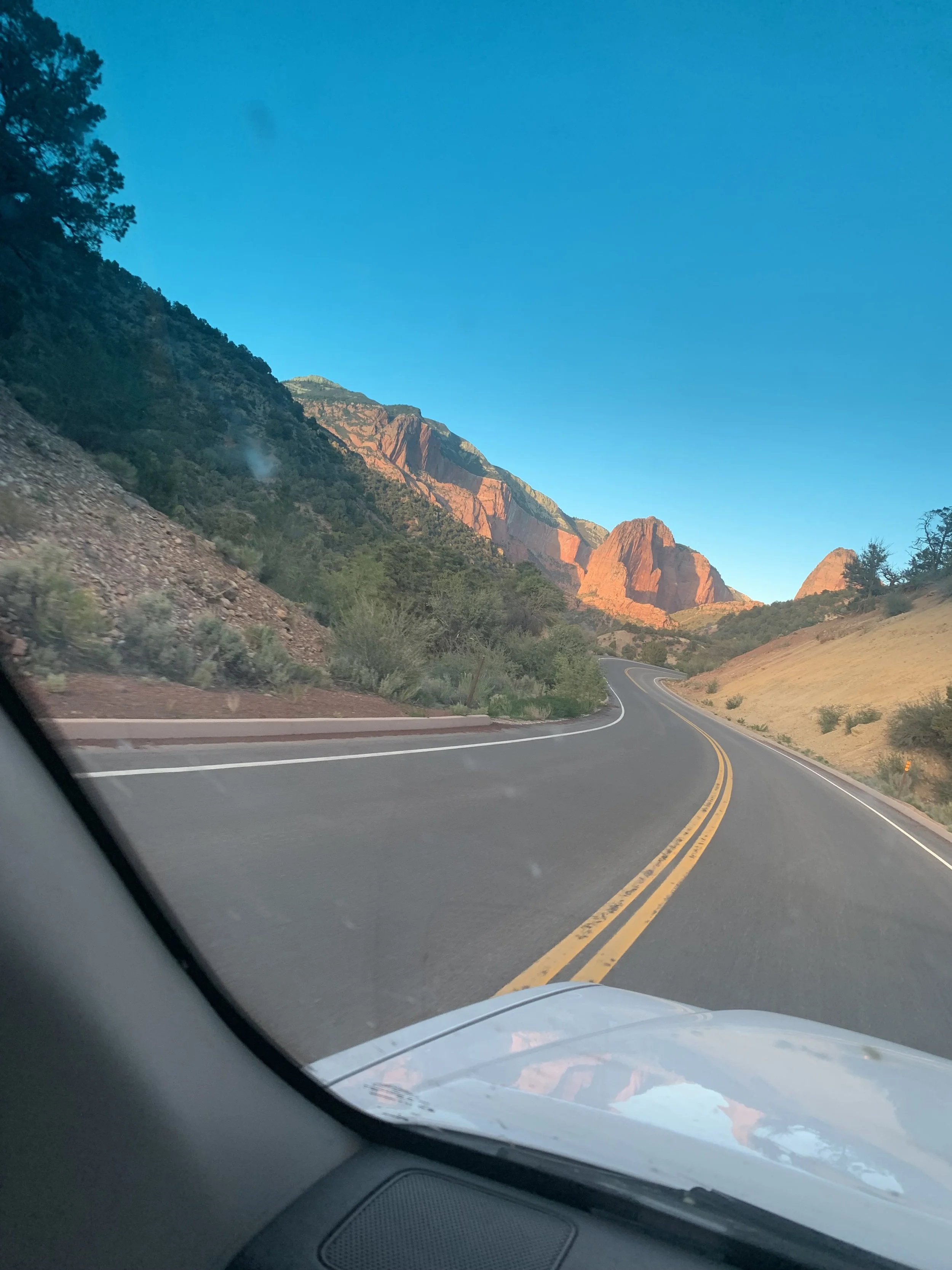 A winding mountain road with a double yellow line, surrounded by rock formations and green foliage under a clear blue sky.