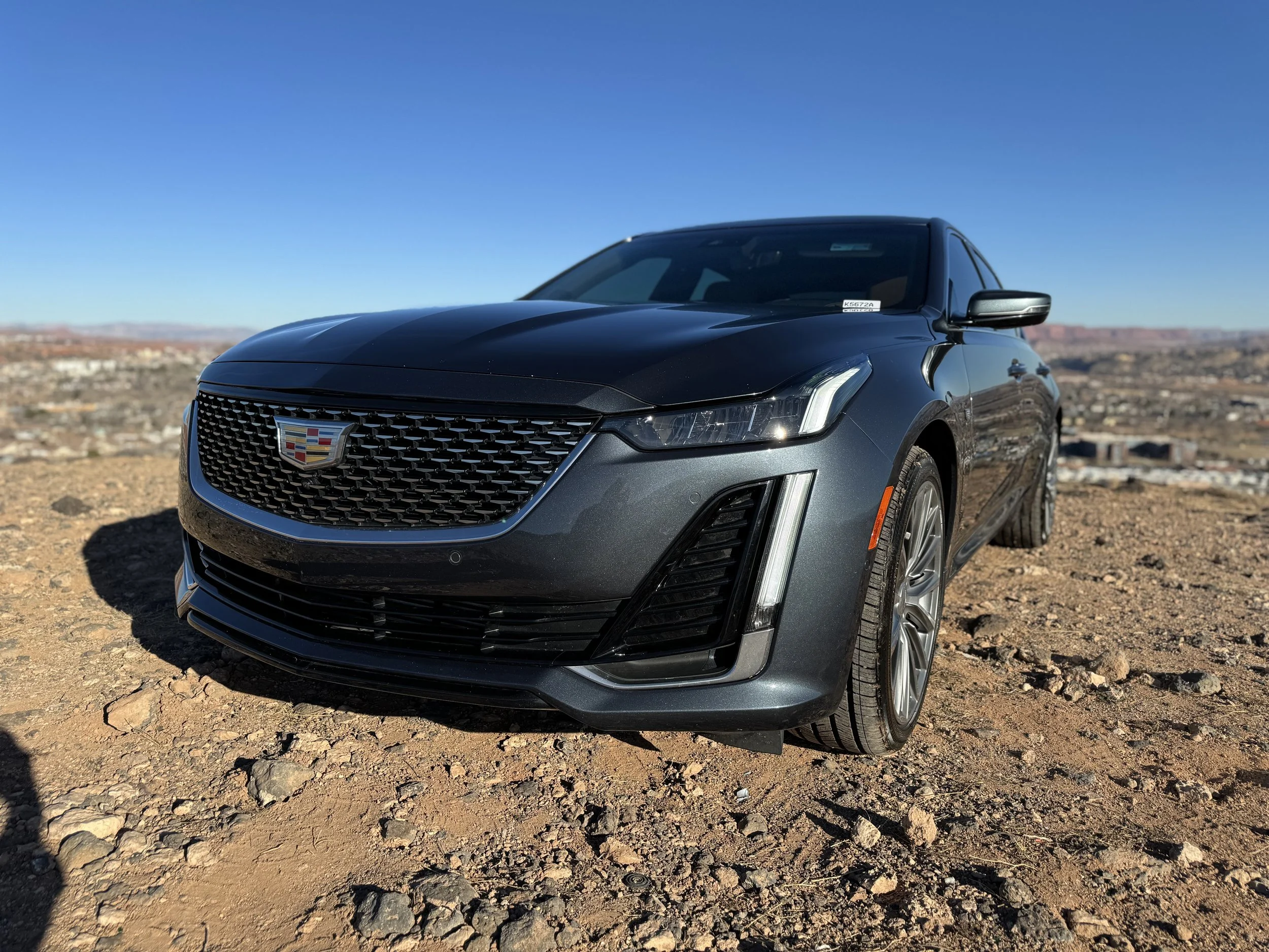 A dark gray Cadillac sedan parked on rocky terrain in a desert landscape with clear blue sky.