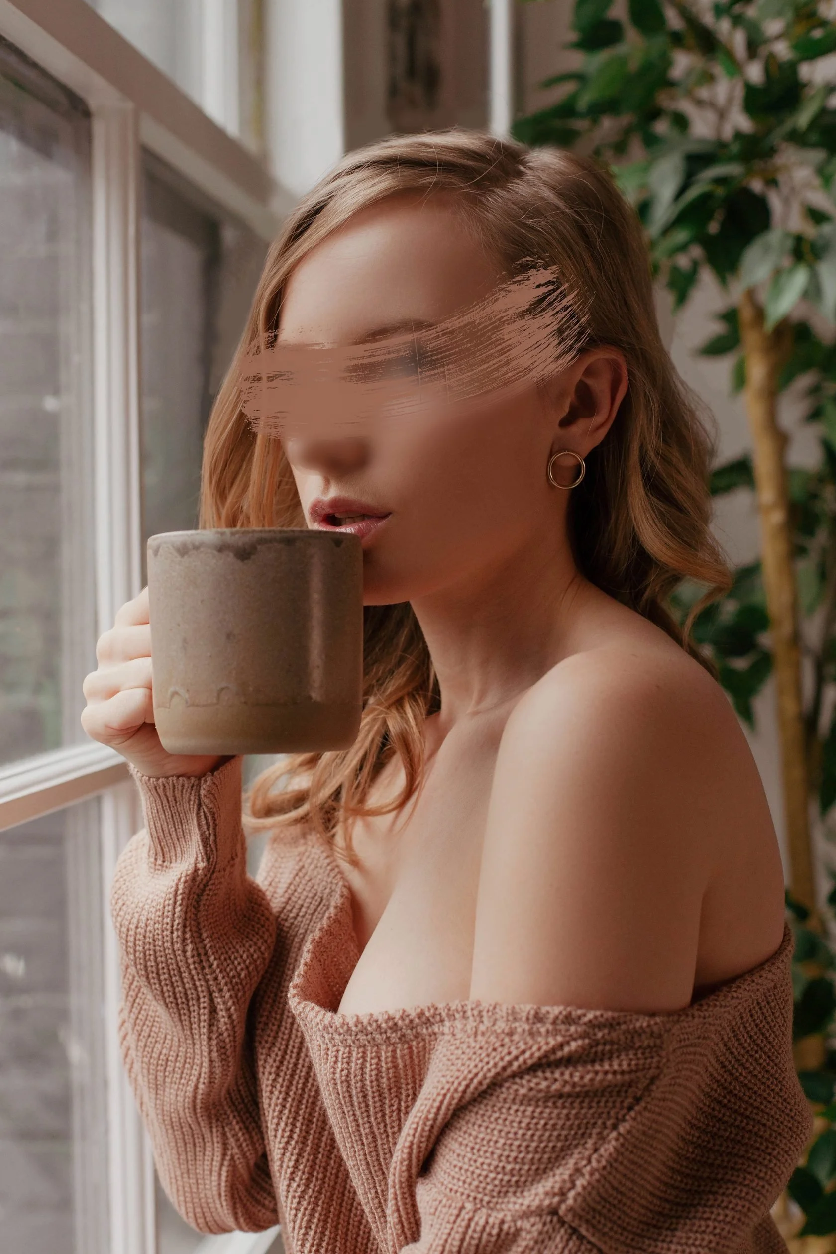 A woman with red hair holding a brown mug near her mouth, wearing a beige off-the-shoulder sweater, standing next to a window with green plants in the background.