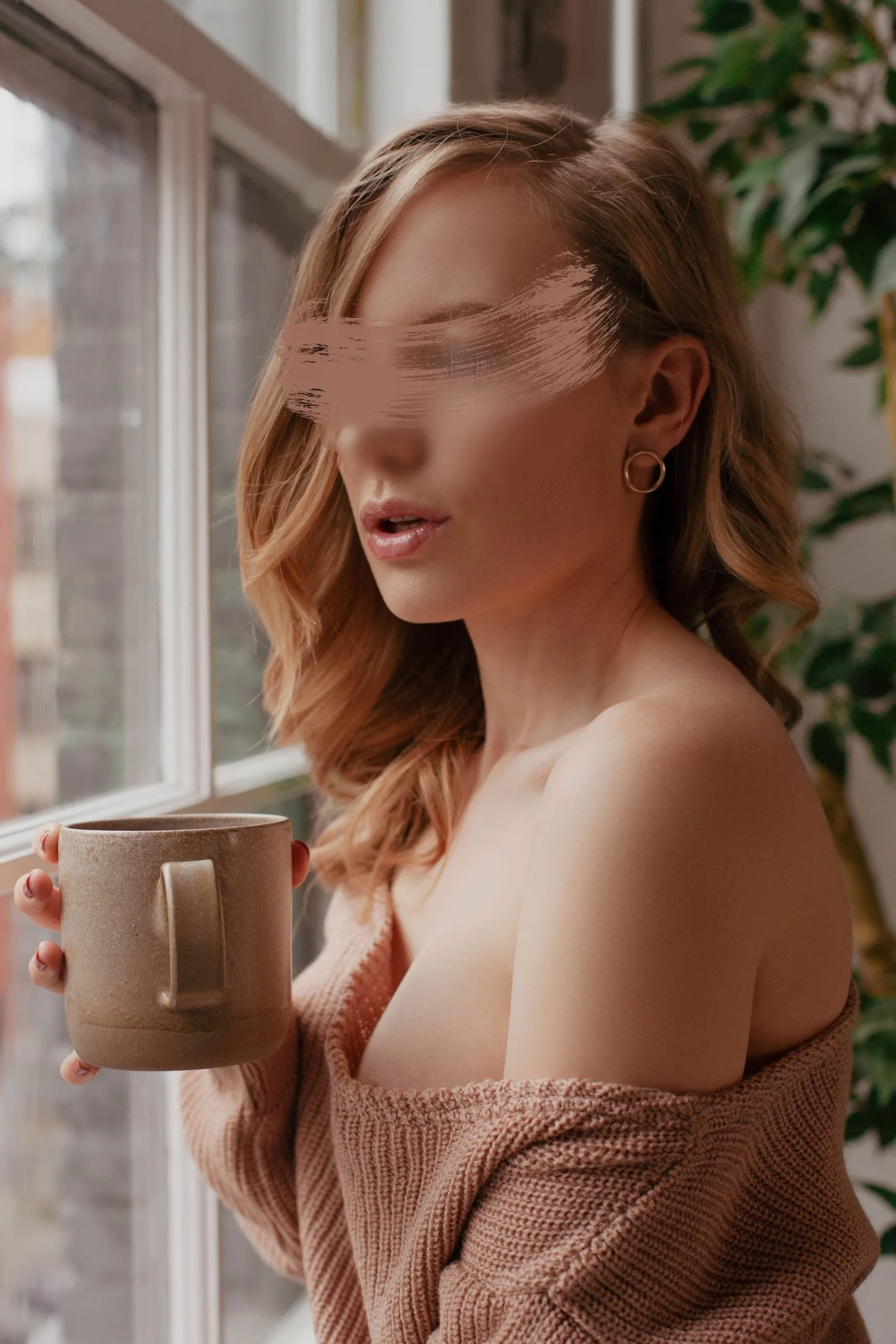 A woman with shoulder-length wavy red hair, wearing gold hoop earrings and a pink off-shoulder sweater, holding a brown ceramic mug while looking out a window.