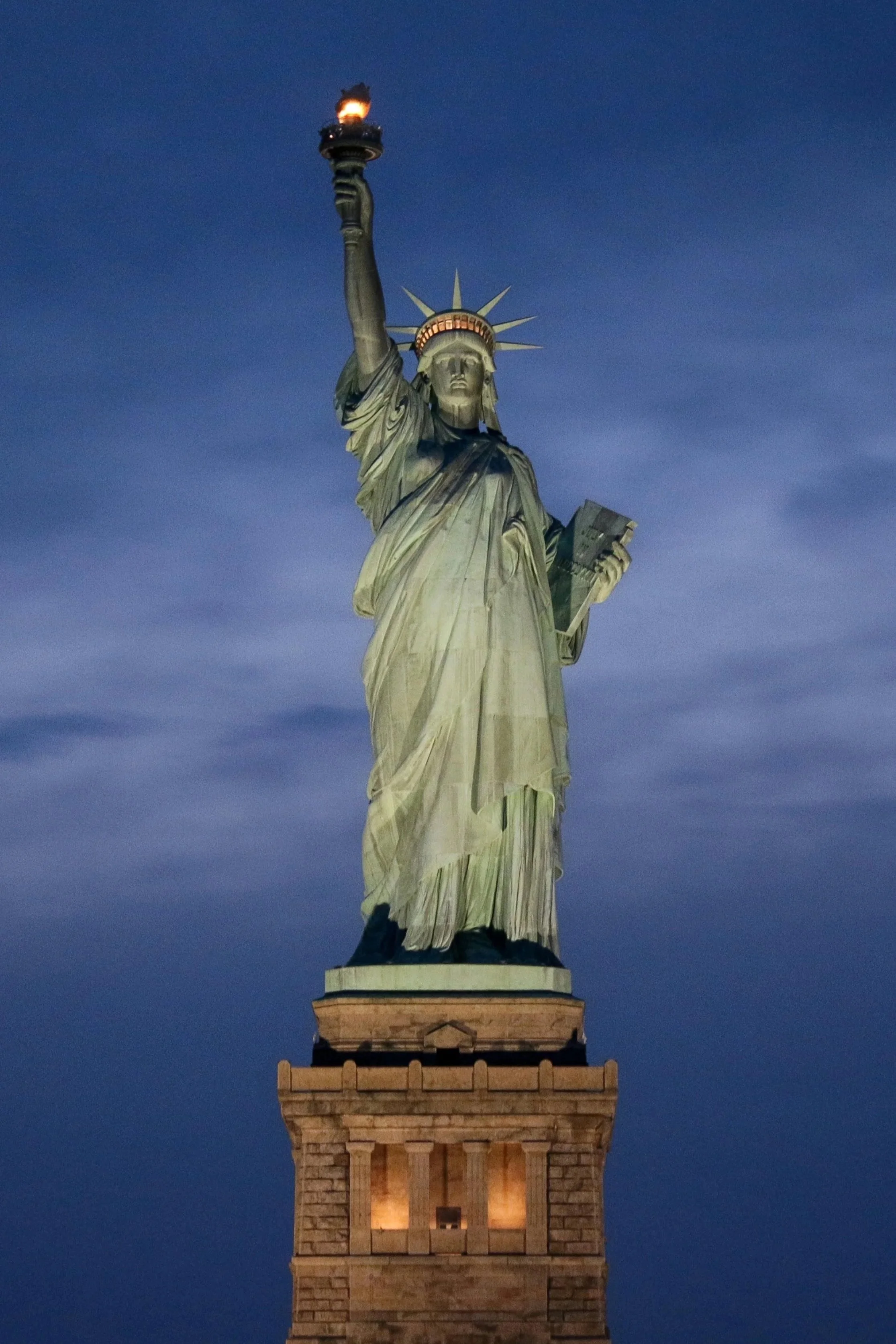 Nighttime image of the Statue of Liberty with a lit torch and bright crown, standing on a stone pedestal against a dark sky.
