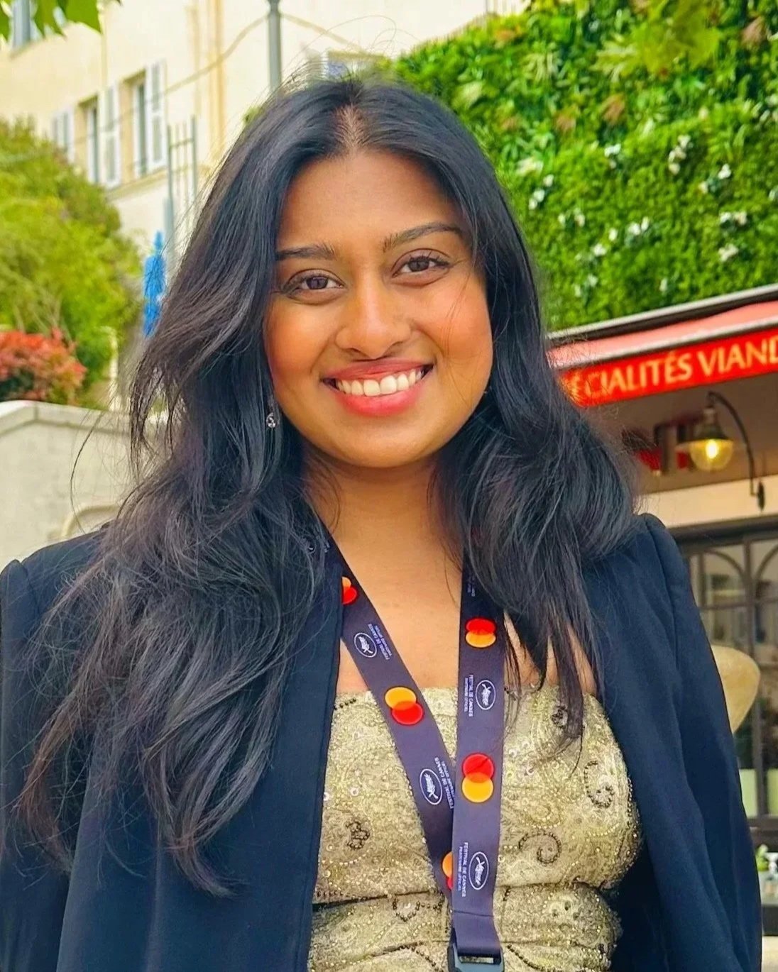 A woman with long black hair smiling outdoors, wearing a dark jacket and a lanyard, with greenery and a building in the background. Meet the founder and CEO of Liminal Frameshift.