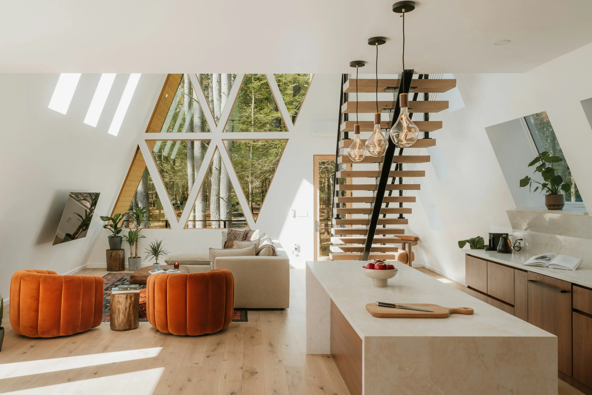 Open-concept living room and kitchen with large triangular windows, modern staircase with hanging light fixtures, beige sofa, orange velvet chairs, potted plants, and wooden cabinets, surrounded by trees visible outside.