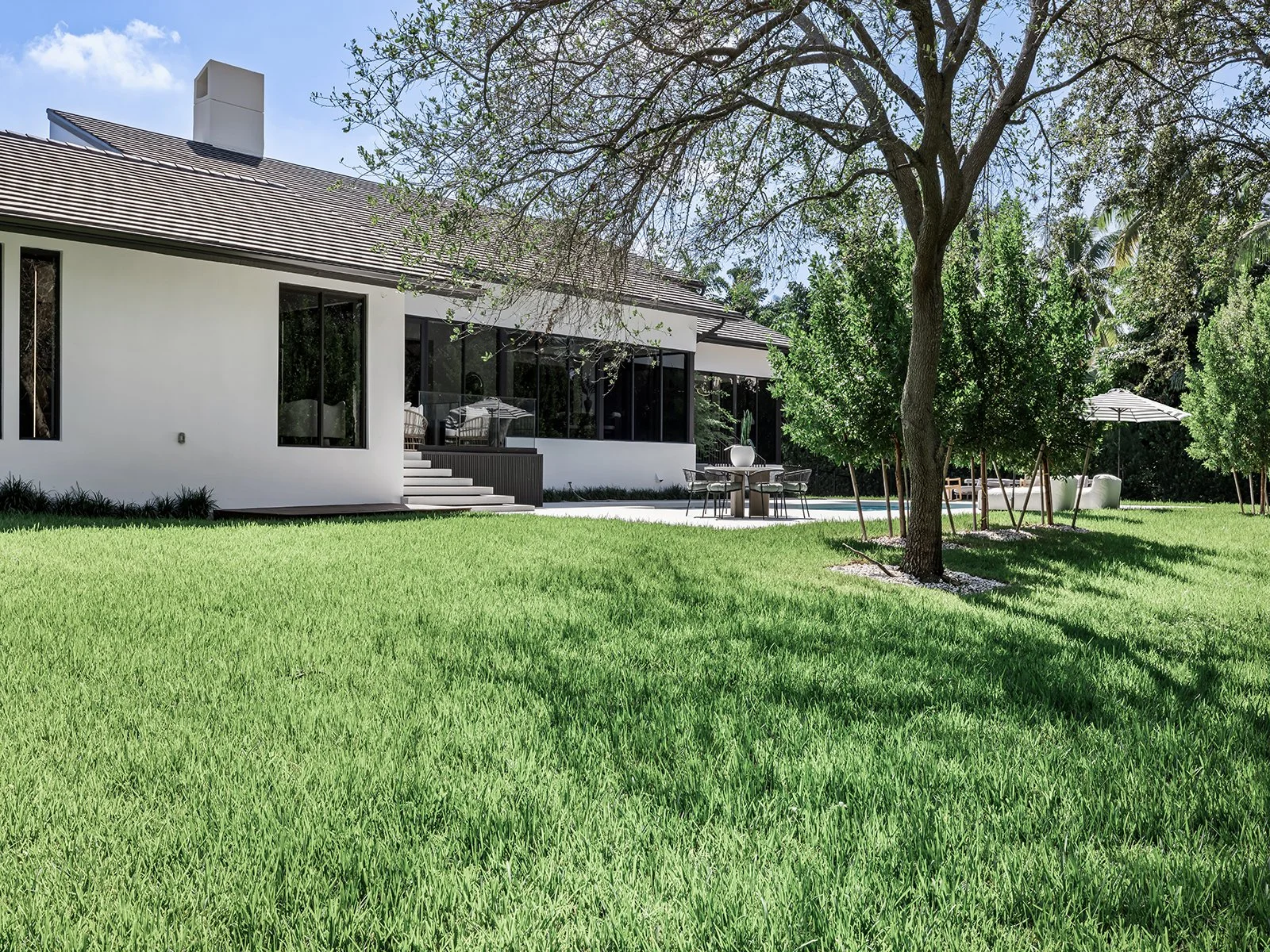 Modern white house with large windows, a lawn, and trees in the backyard with patio furniture and umbrellas.