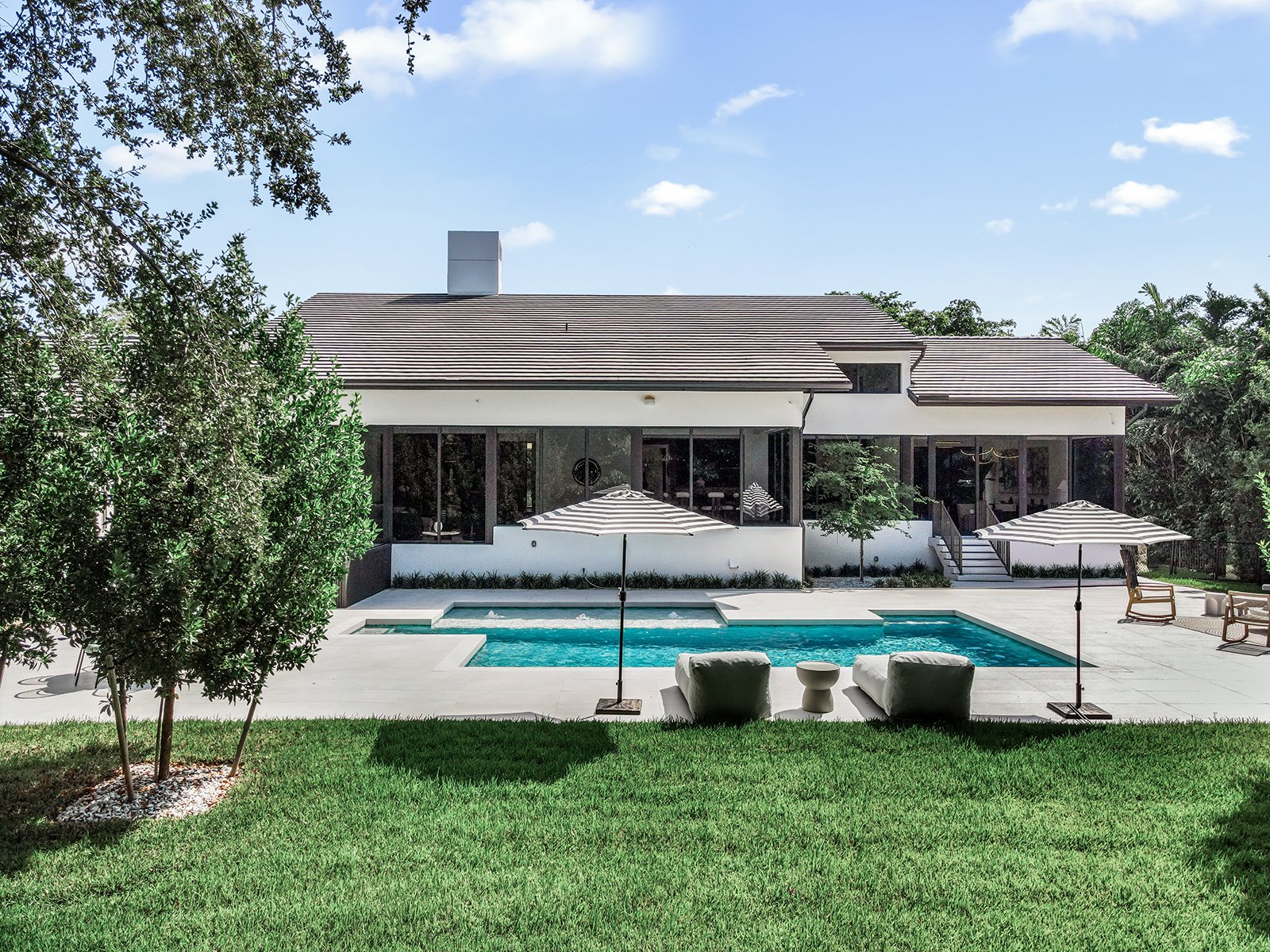 Modern house with large windows, backyard pool, umbrellas, lounge chairs, and green lawn on a sunny day.
