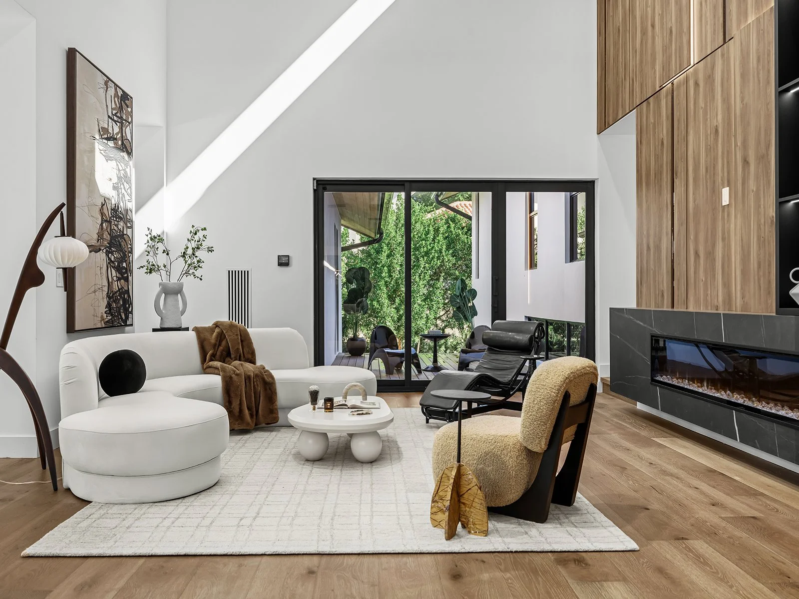 Living room with white sofa, black chairs, cozy armchair, modern fireplace, wooden wall panels, sliding glass door opening to balcony with green trees, wooden floor, abstract artwork, and decorative items.