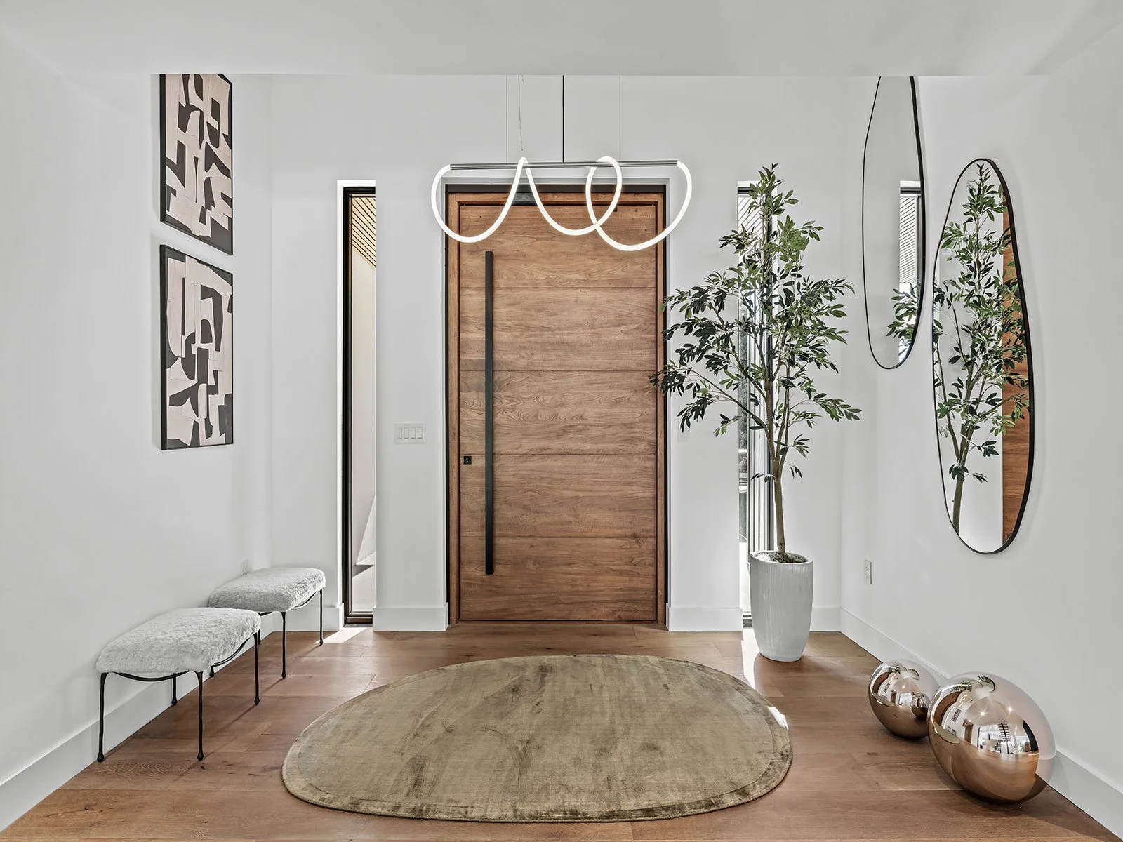 Modern entryway with wooden door, white walls, abstract wall art, a large potted tree, oval mirrors, a beige oval rug, and metallic decorative spheres on a hardwood floor.