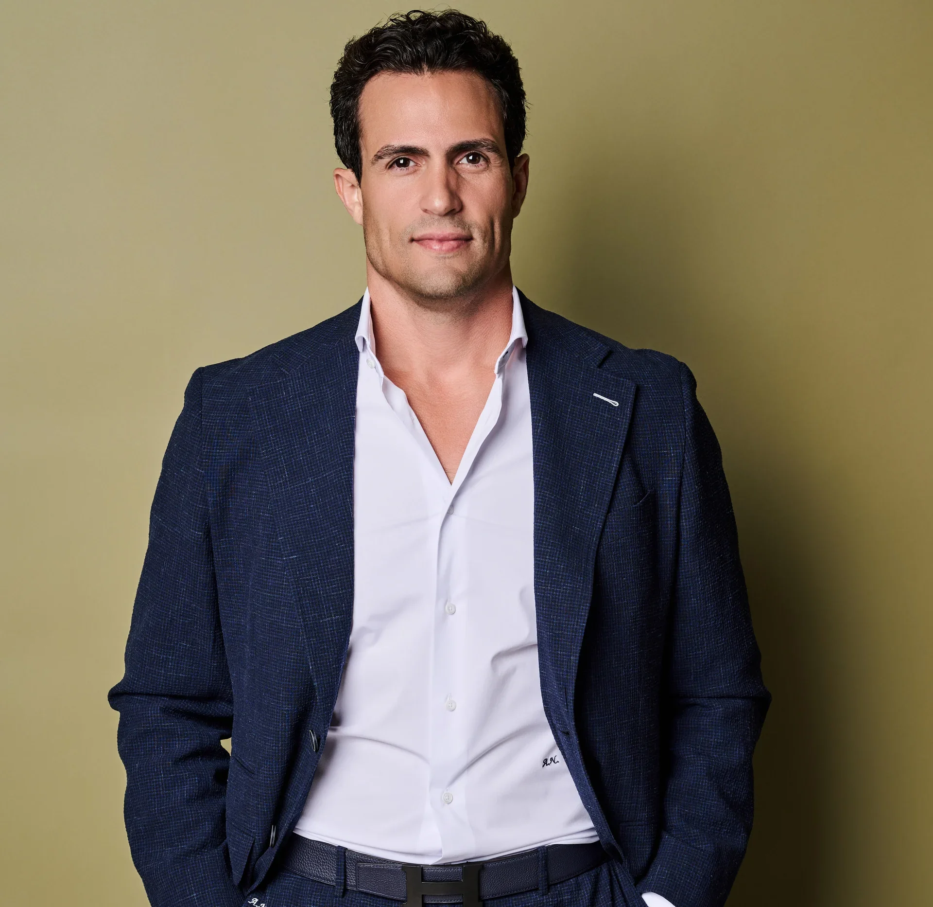 A man with dark curly hair in a blue suit and white shirt standing against a yellow background.