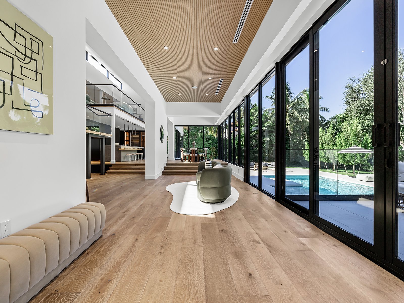 Spacious modern living room with large glass sliding doors showing an outdoor pool and lush greenery, wooden flooring, and minimalist furniture.
