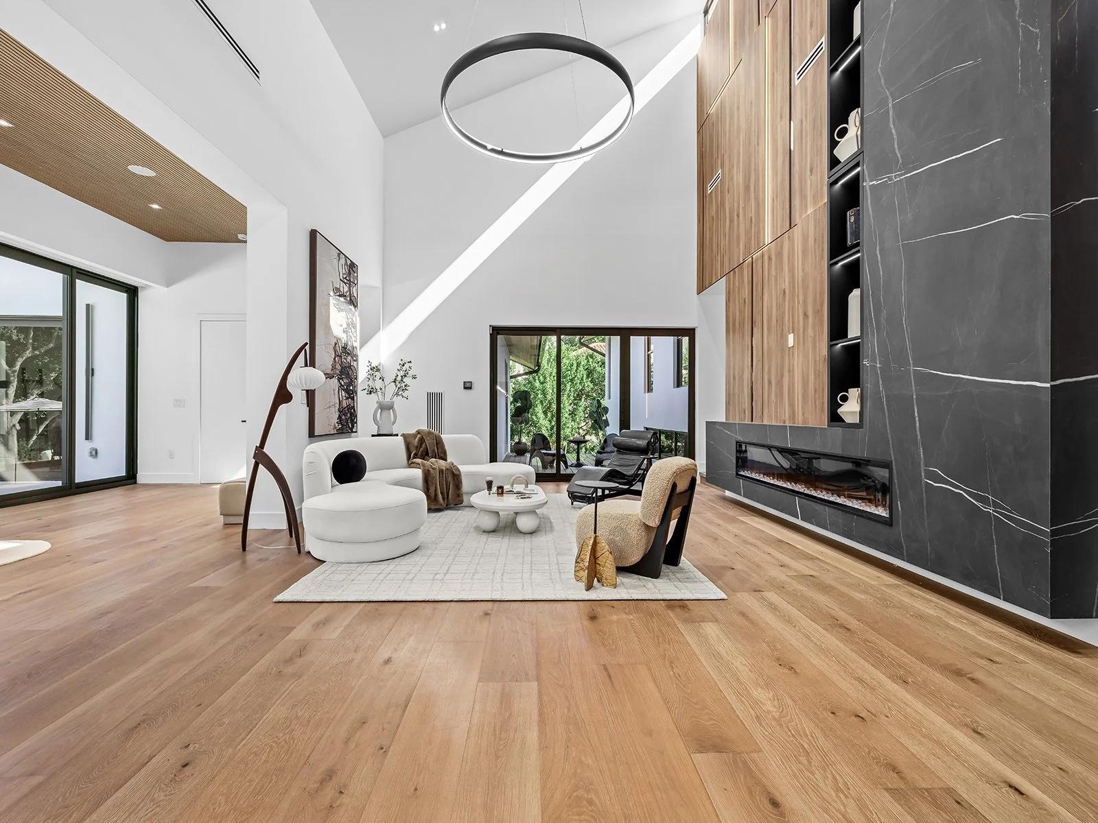 Modern living room with white walls, a vaulted ceiling, and a large circular chandelier. Features a mix of furniture including a curved white sofa, black and beige chairs, and a round coffee table on a light-colored rug. There is a fireplace with bla