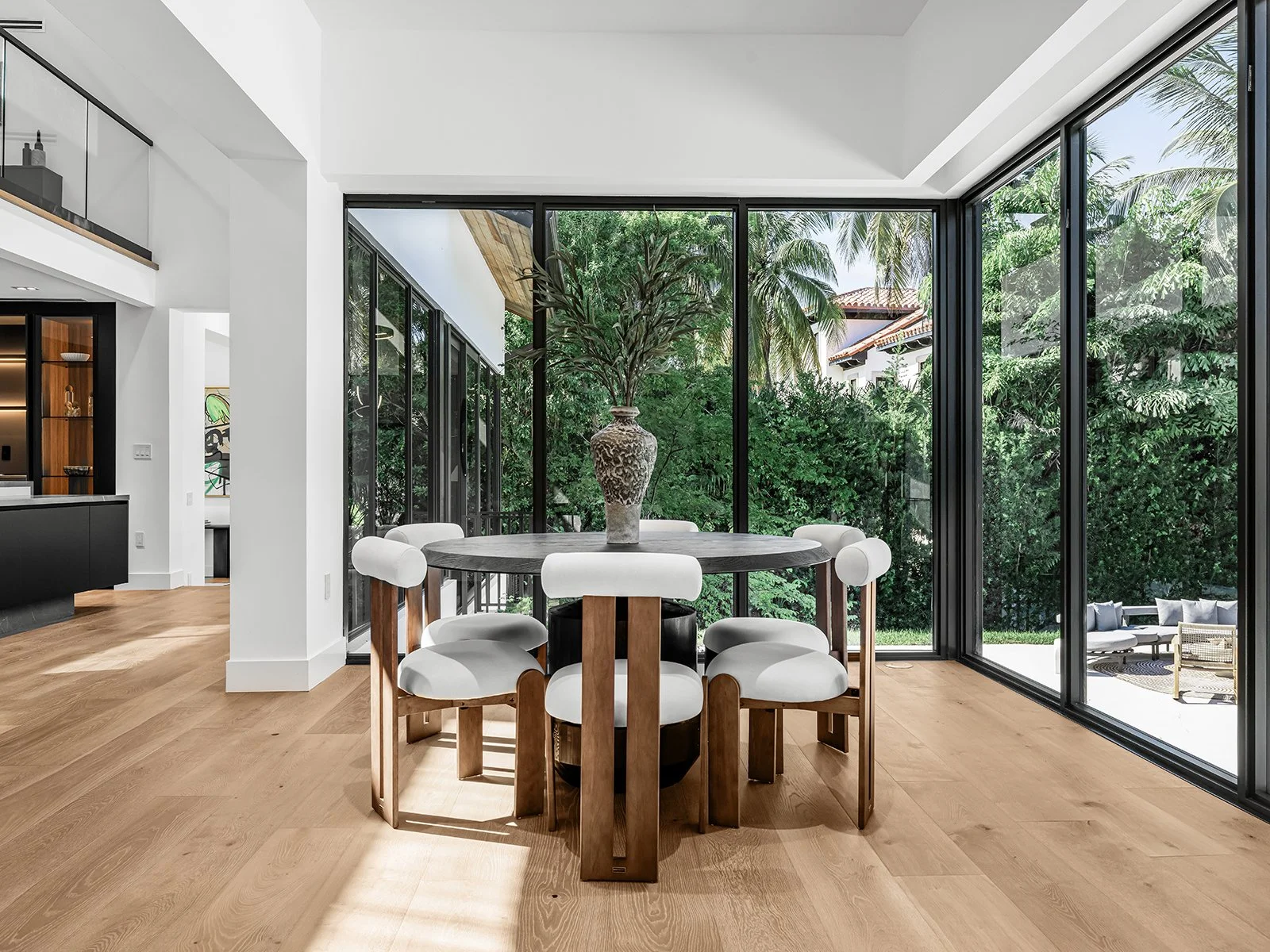 Modern dining room with round wooden table, white cushioned chairs, large vases, and floor-to-ceiling windows showing greenery outside.