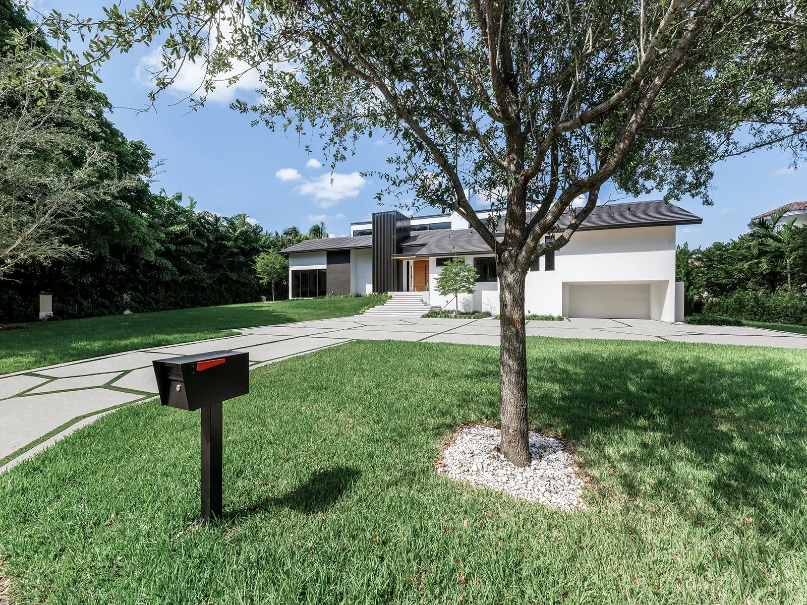 Modern white house with gray roof, driveway, and garage, surrounded by green lawn and trees, with a mailbox in the foreground.
