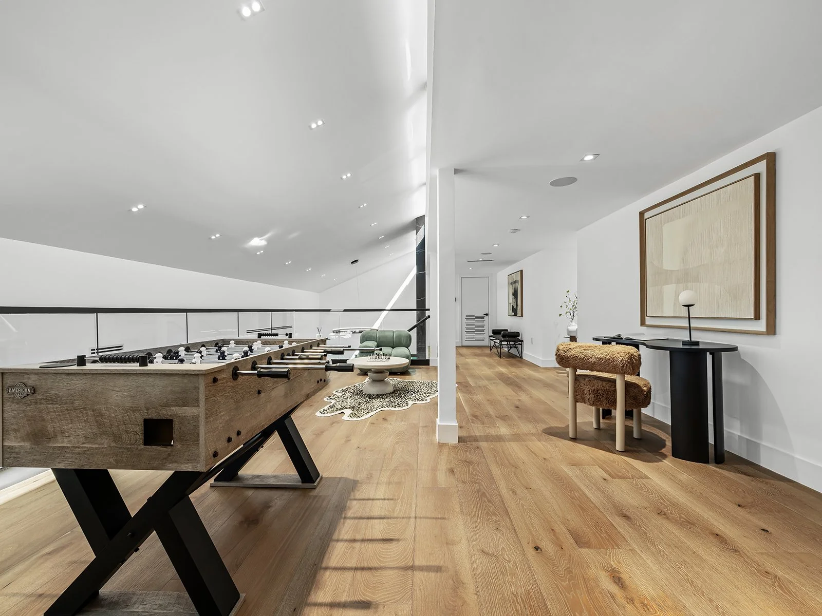 Modern attic living space with foosball table, green armchair, decorative rug, minimalist art, round side table, and cozy fur stool. Hardwood floors and white walls with recessed lighting.
