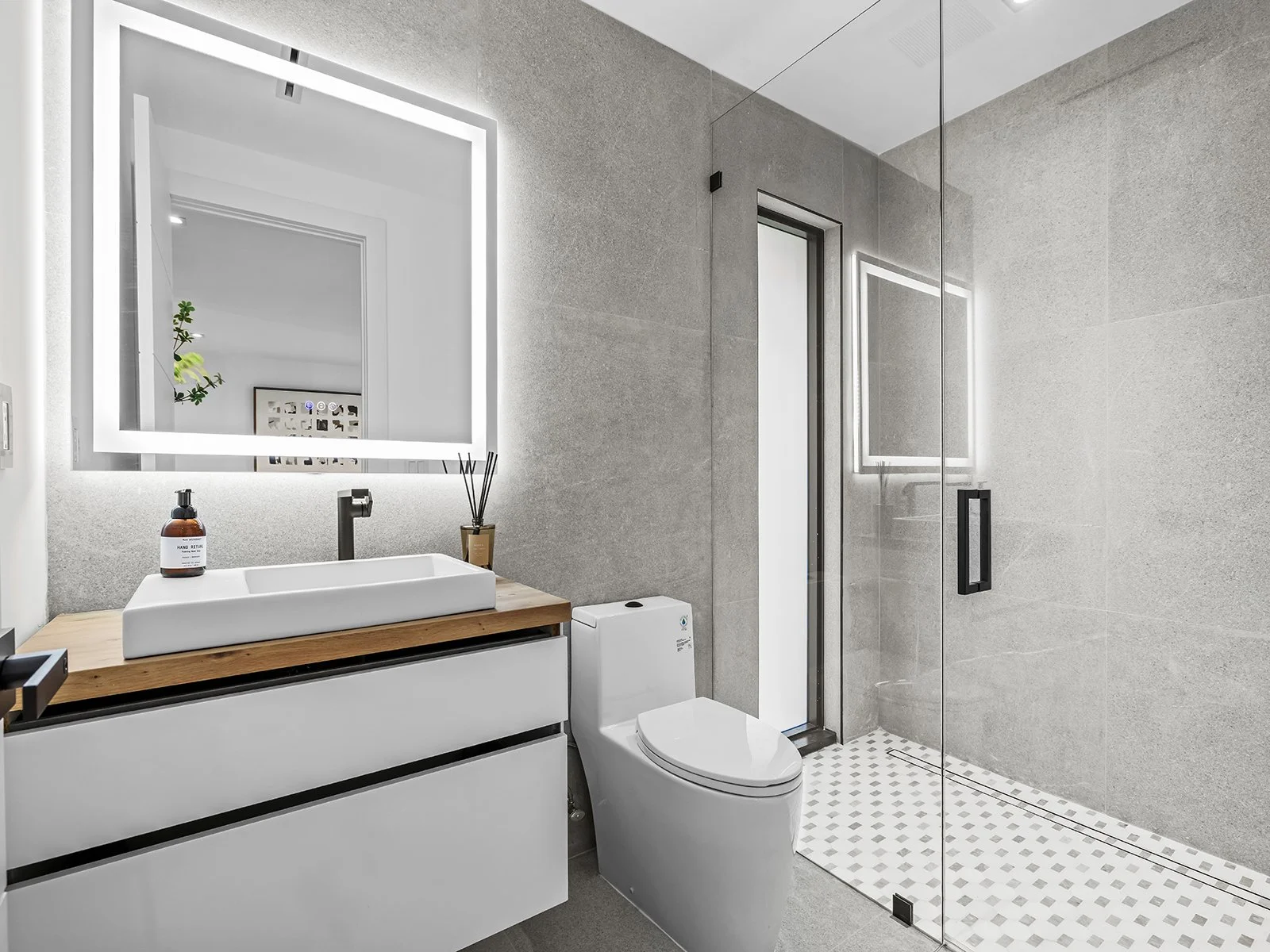 Modern bathroom with gray tiled walls, a glass shower enclosure, a white toilet, and a white vanity with a vessel sink and wooden countertop, illuminated by a backlit mirror.