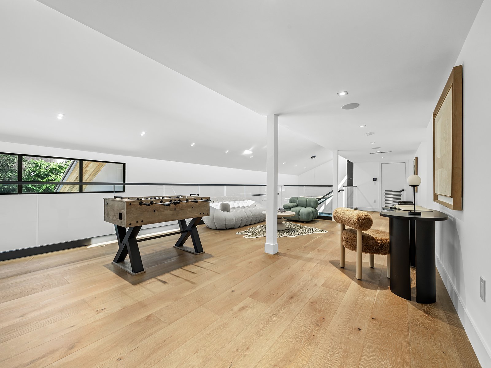 Spacious modern loft with wooden flooring, foosball table, white sofa, green armchair, eclectic decor, and large windows letting in natural light.