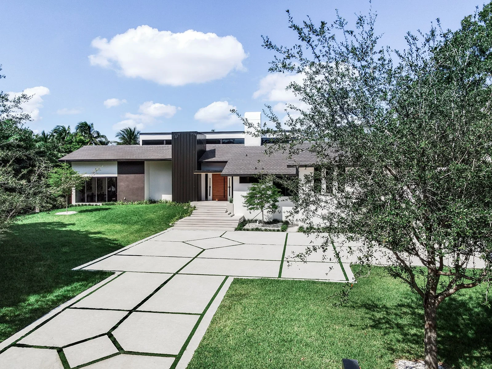 Modern house with gray roof, black vertical siding, and white exterior walls, surrounded by a green lawn, trees, and a geometric concrete driveway, under a blue sky with white clouds.