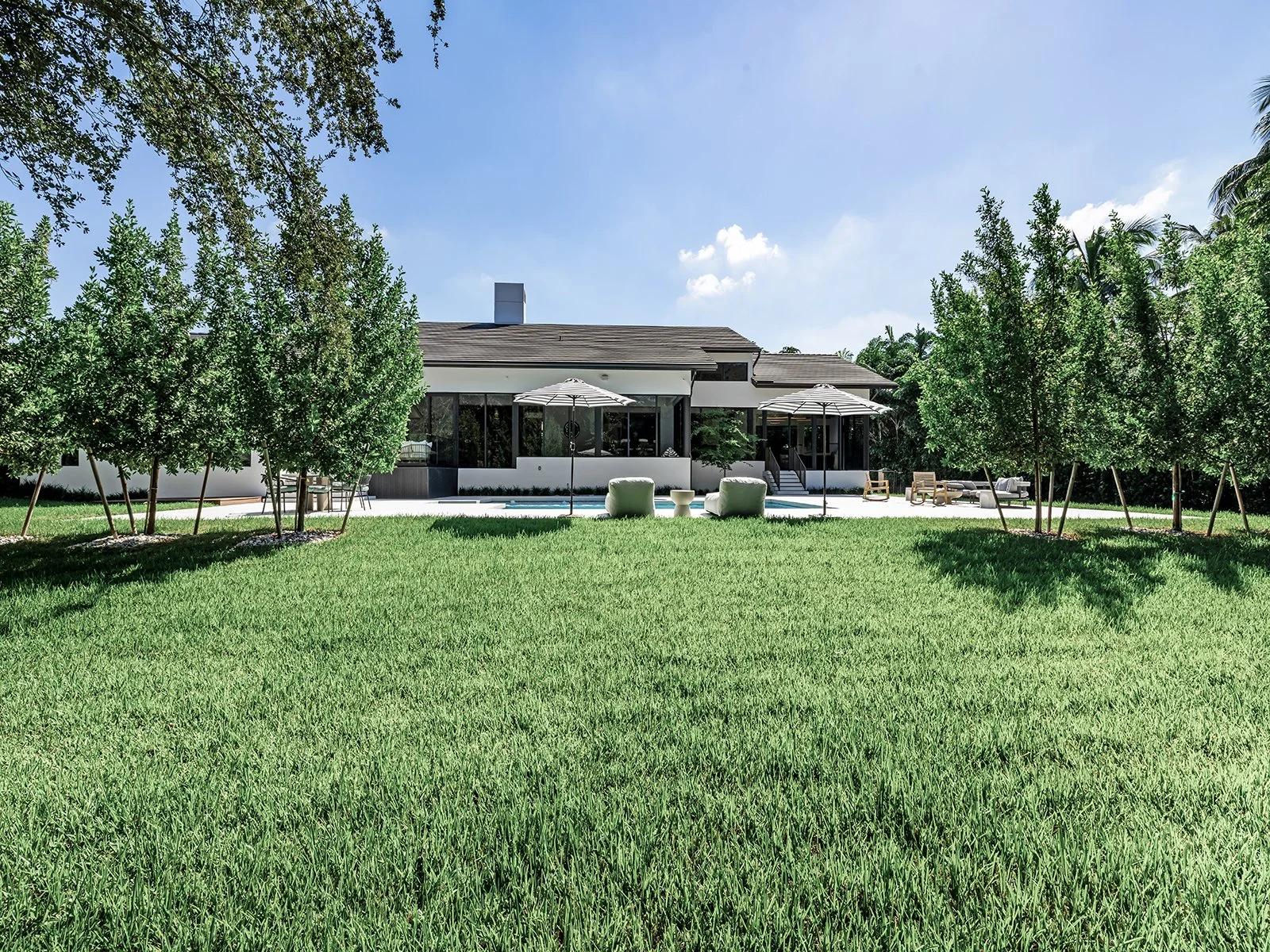 Modern house with a backyard patio, surrounded by trees, with outdoor chairs, umbrellas, and a green lawn.
