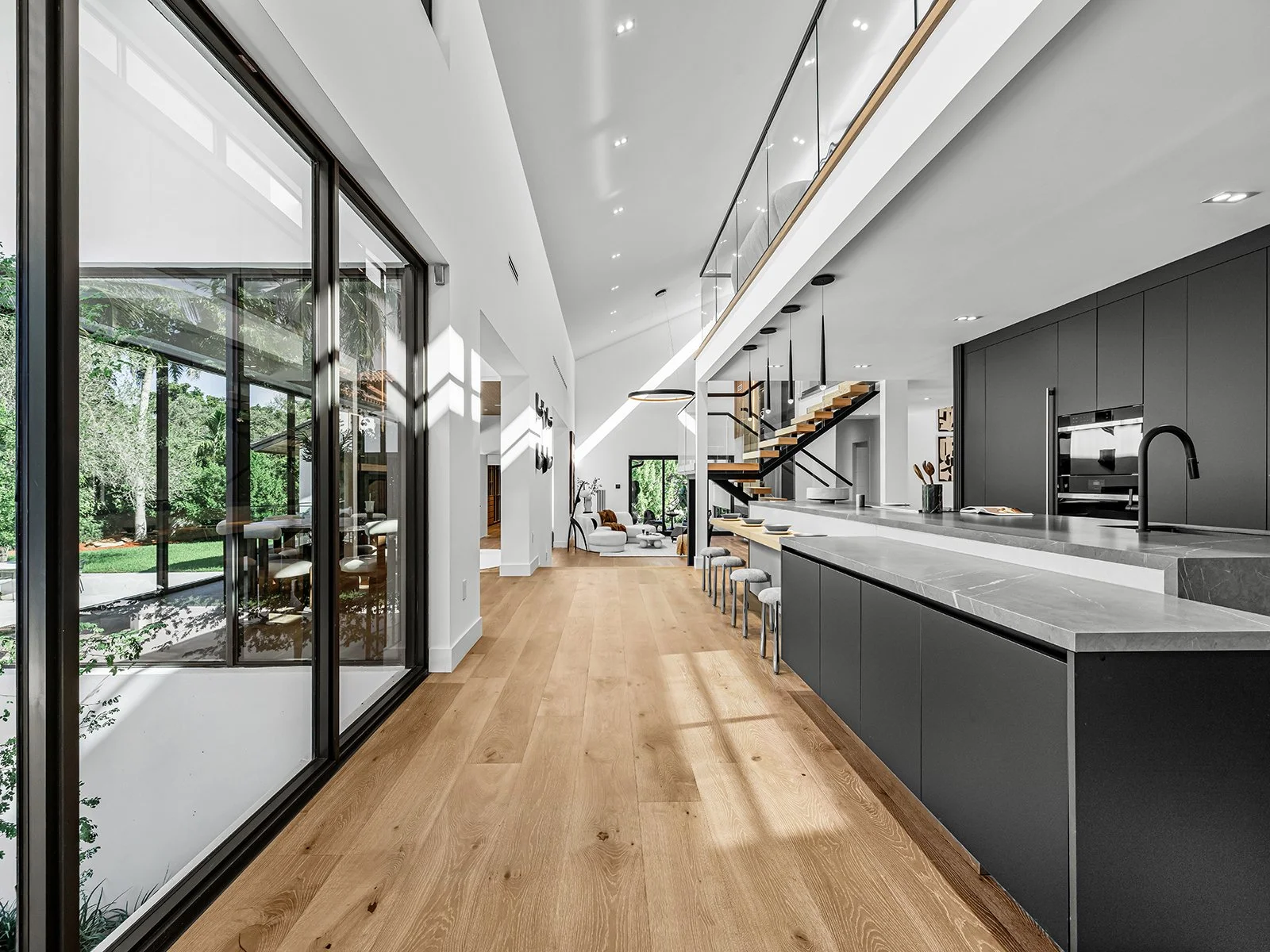 Modern kitchen and living area with large windows, black cabinets, marble countertops, a staircase, and a view of a backyard with trees.
