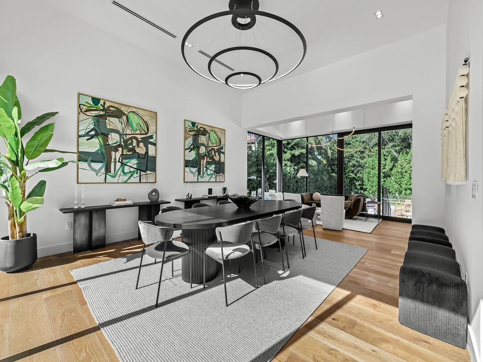Modern dining and living room with white walls, wood flooring, black dining table, gray chairs, black sideboard, colorful abstract art, green potted plant, large windows with greenery outside, and seating area with armchair and sofa.