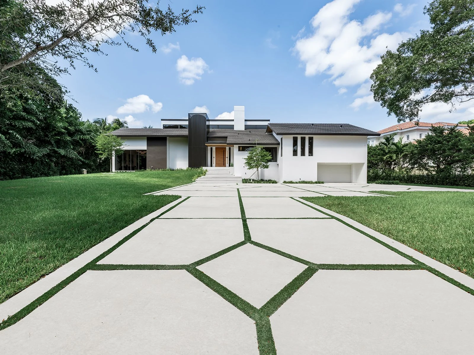 Modern white house with a landscaped yard and a concrete driveway leading to the entrance, surrounded by lush green trees under a blue sky with scattered clouds.