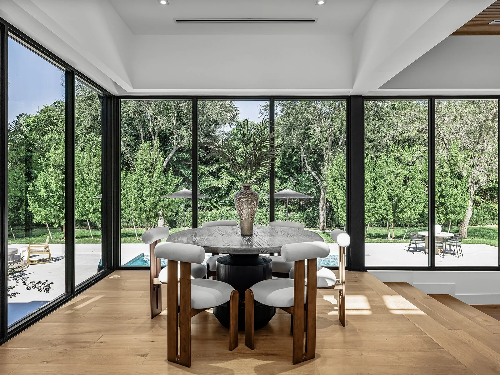 Modern dining room with large glass windows overlooking a backyard with trees, outdoor seating, and a pool. Circular dining table with a decorative vase and white chairs.