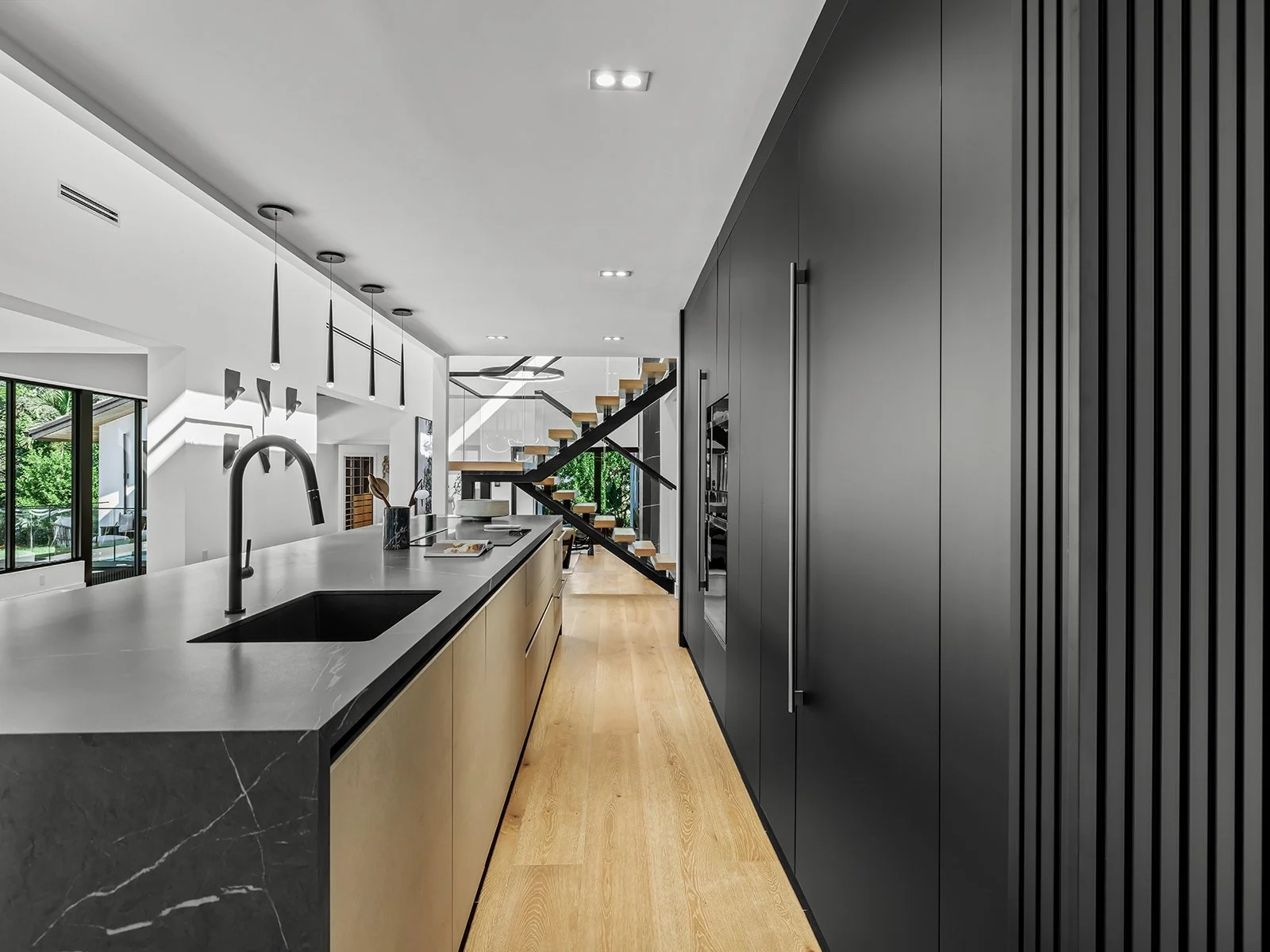 Modern kitchen with black and beige cabinetry, white countertop with black sink, black faucet, and pendant lights. Open floor plan with staircase visible in background.