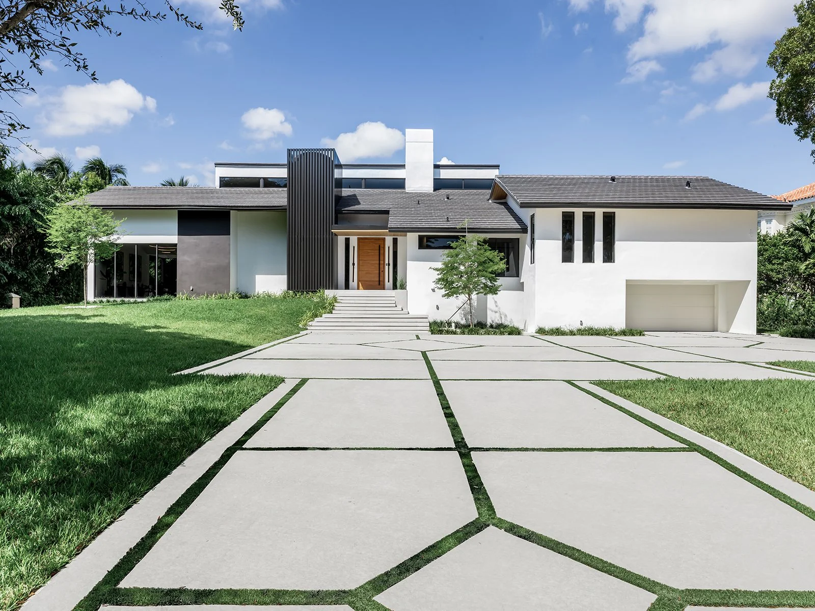 Modern house with a white exterior, a gray roof, and a large front yard with a concrete pathway leading to the entrance. The house features large windows, a wooden front door, and landscaped greenery.