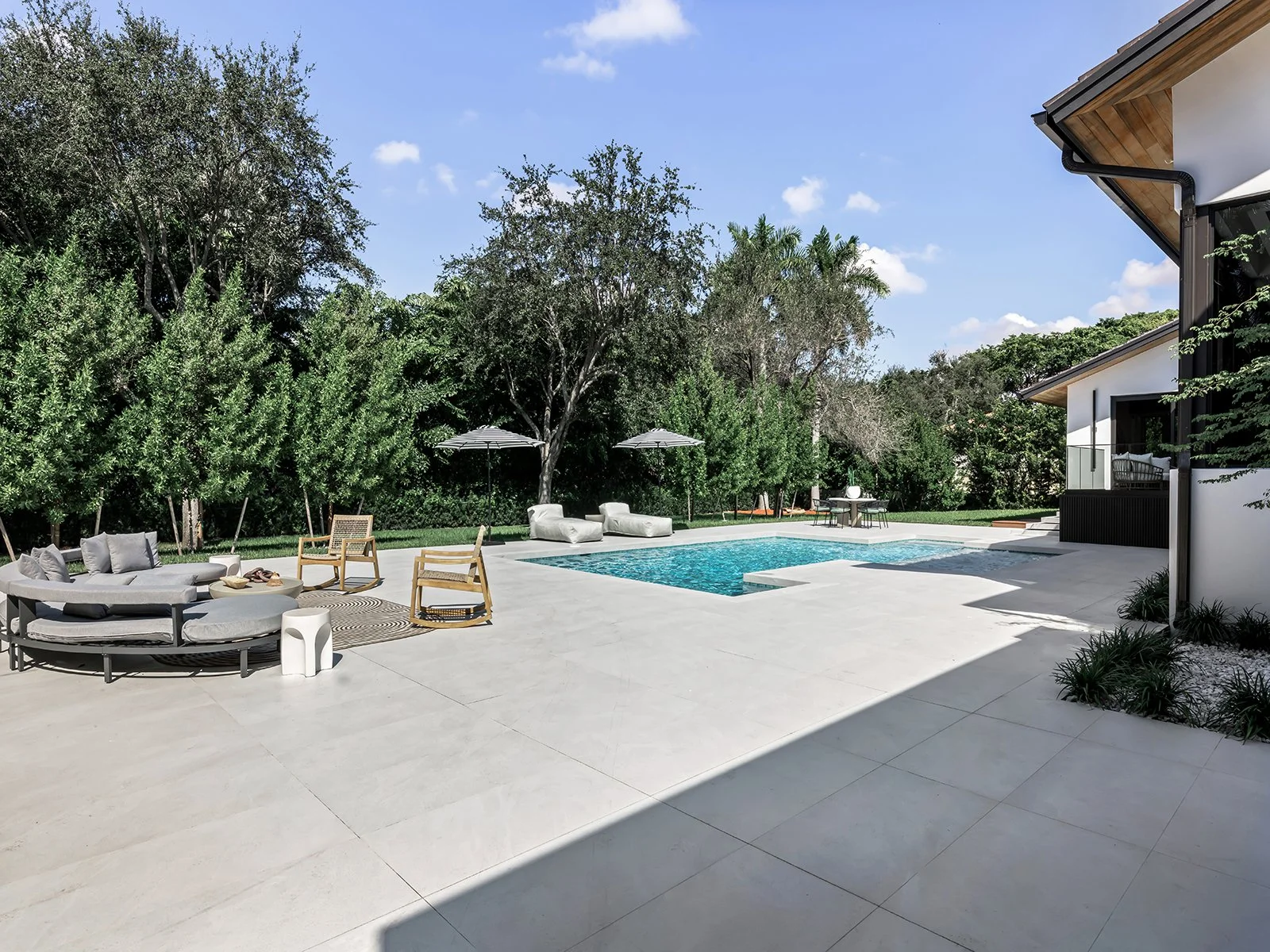 Modern backyard with a swimming pool, outdoor seating with cushions, and an umbrella, surrounded by trees, adjacent to a contemporary house with a balcony and a white exterior.