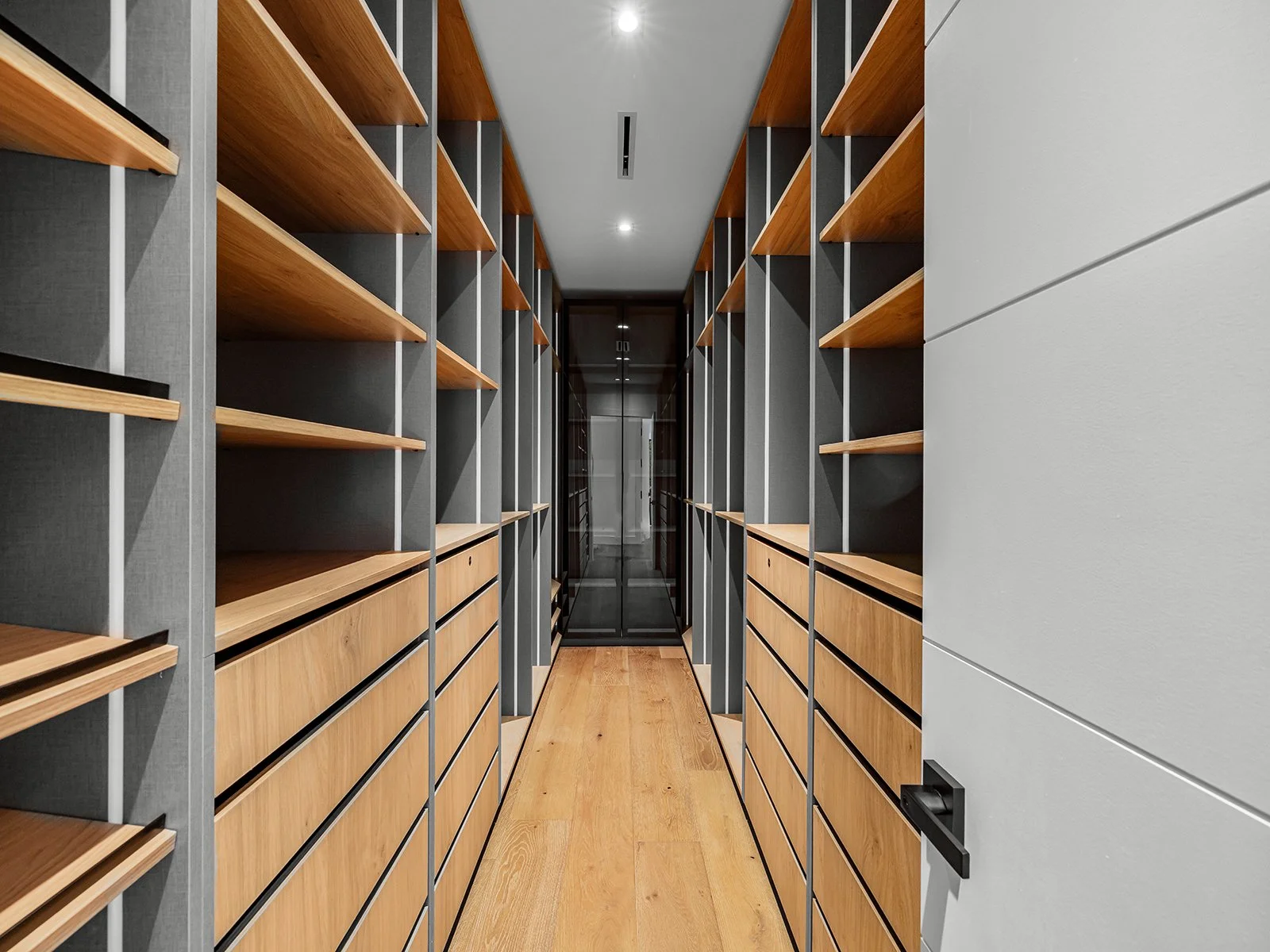A modern walk-in closet with wooden shelves and drawers on both sides, a wooden floor, and a glass door at the end.