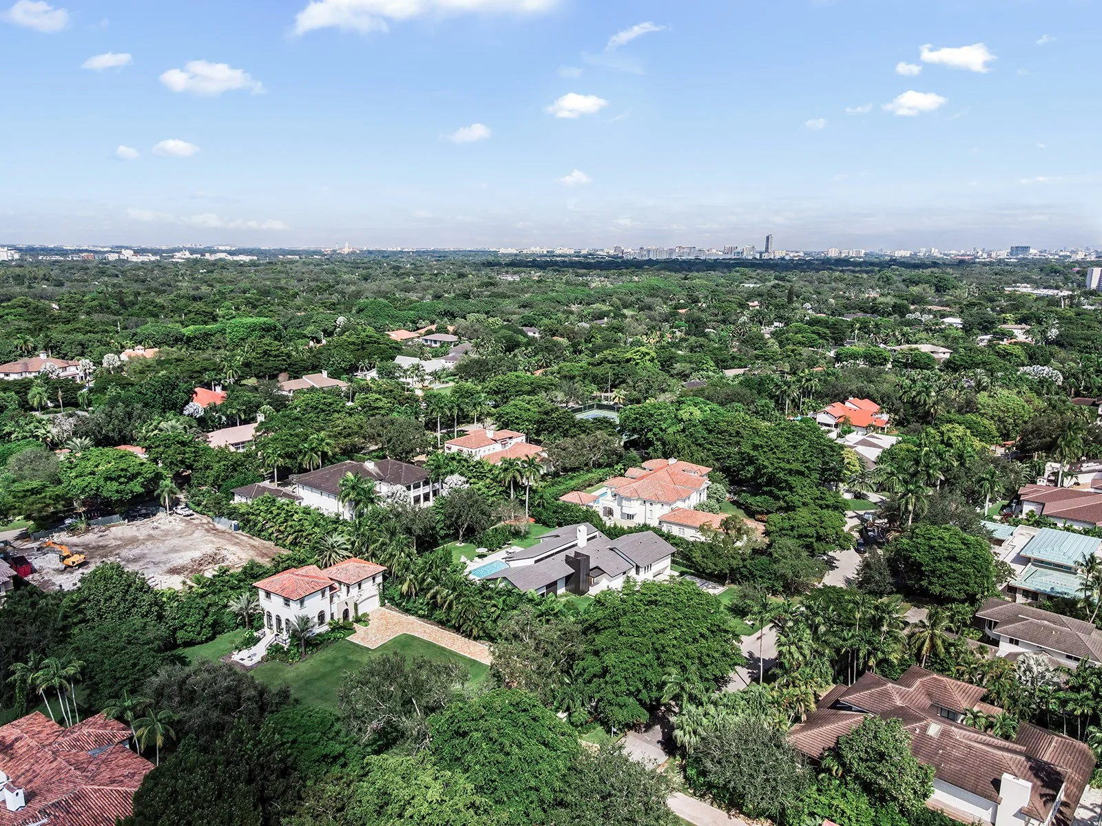 Aerial view of a residential neighborhood with many trees and houses, some with red-tiled roofs, under a bright blue sky with scattered clouds.