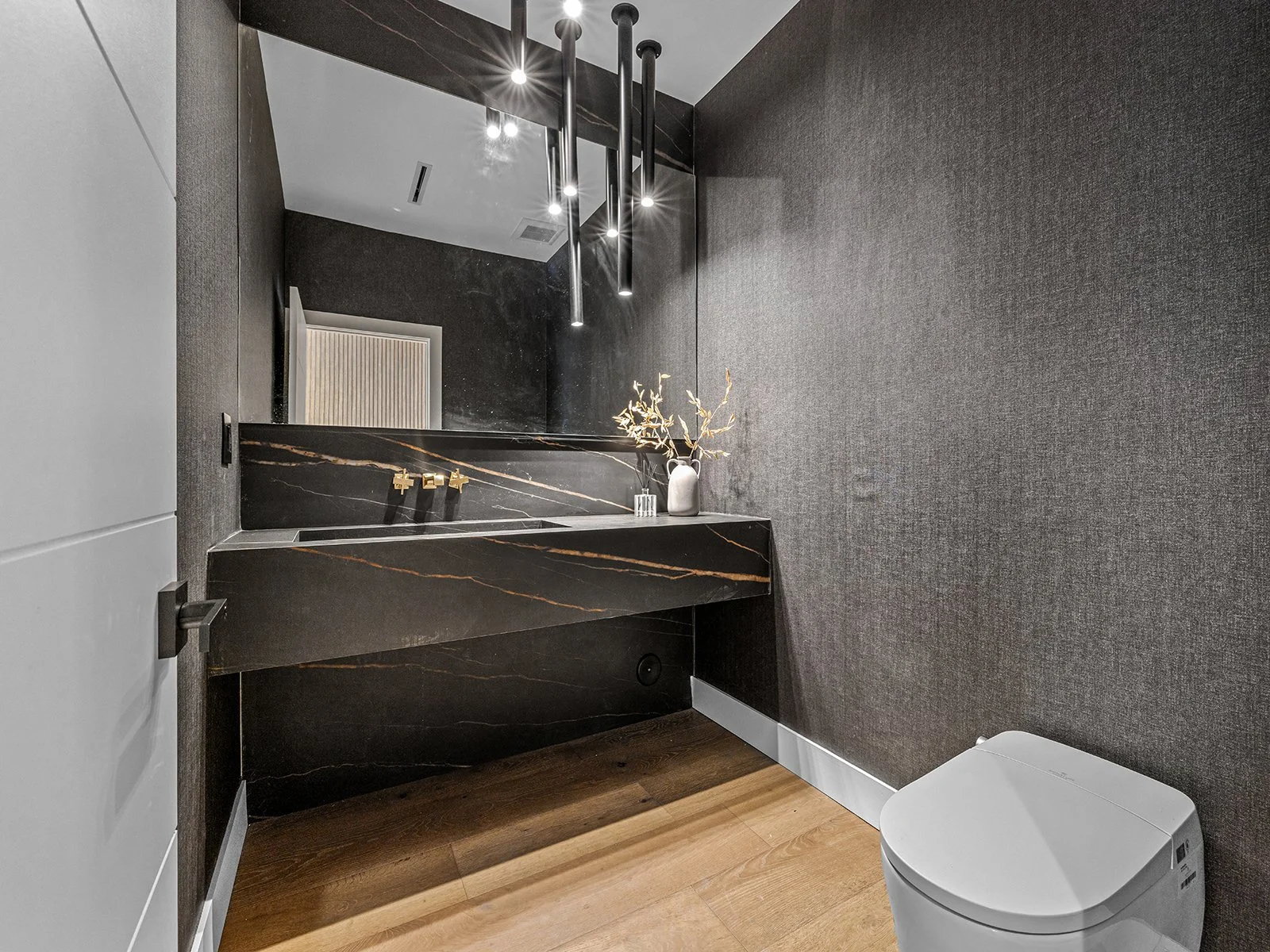 Modern bathroom with dark textured walls, wooden floor, black marble sink with gold fixtures, large mirror with black pendant lights, white toilet, and a decorative white vase with dried branches.