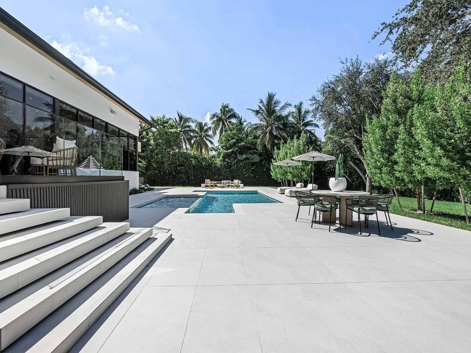 Modern backyard with swimming pool, outdoor seating, and lush tropical trees in a sunny setting.