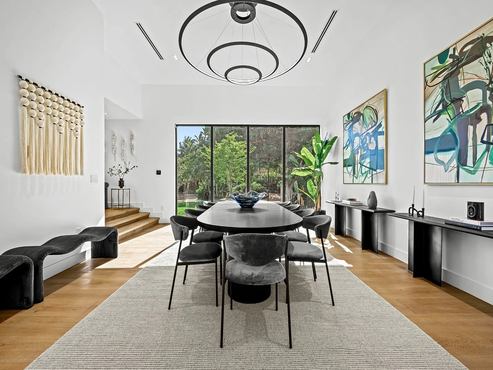 Modern dining room with a large black dining table and nine black upholstered chairs, a wood floor, and large glass window showing greenery outside. Artwork on the white walls, and a black console table with decorative items. A circular chandelier ha