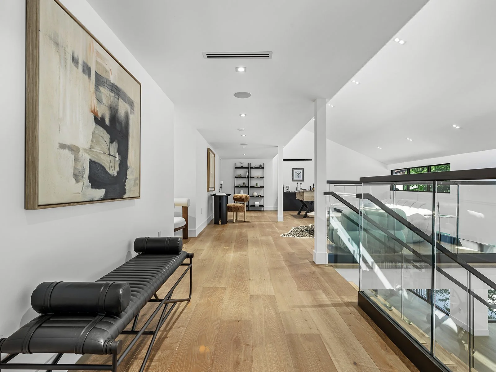 Interior of a modern, minimalist home with white walls, wooden floors, and contemporary furniture. Features a black leather bench, abstract artwork, open stairs with glass railing, and scattered decor items in the background.