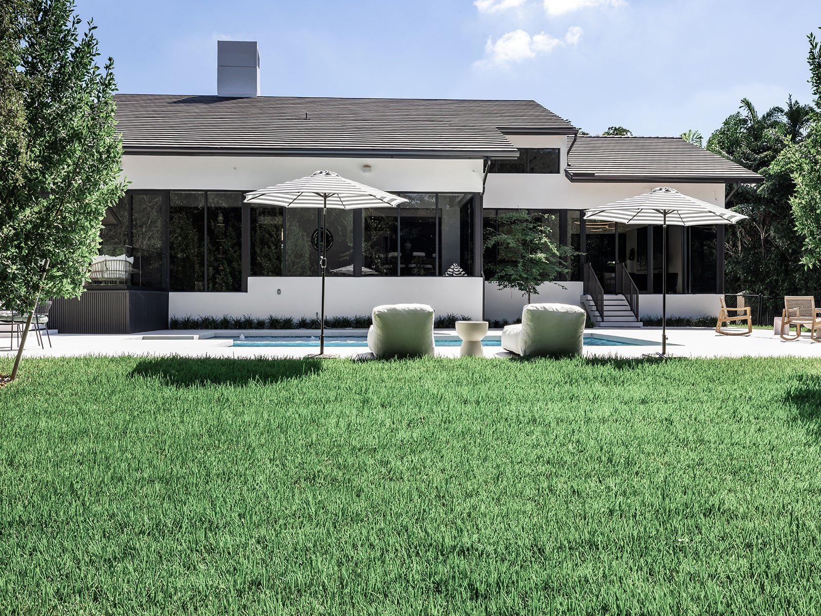 Modern house with a backyard pool area featuring two striped umbrellas, lounge chairs, and lush greenery.