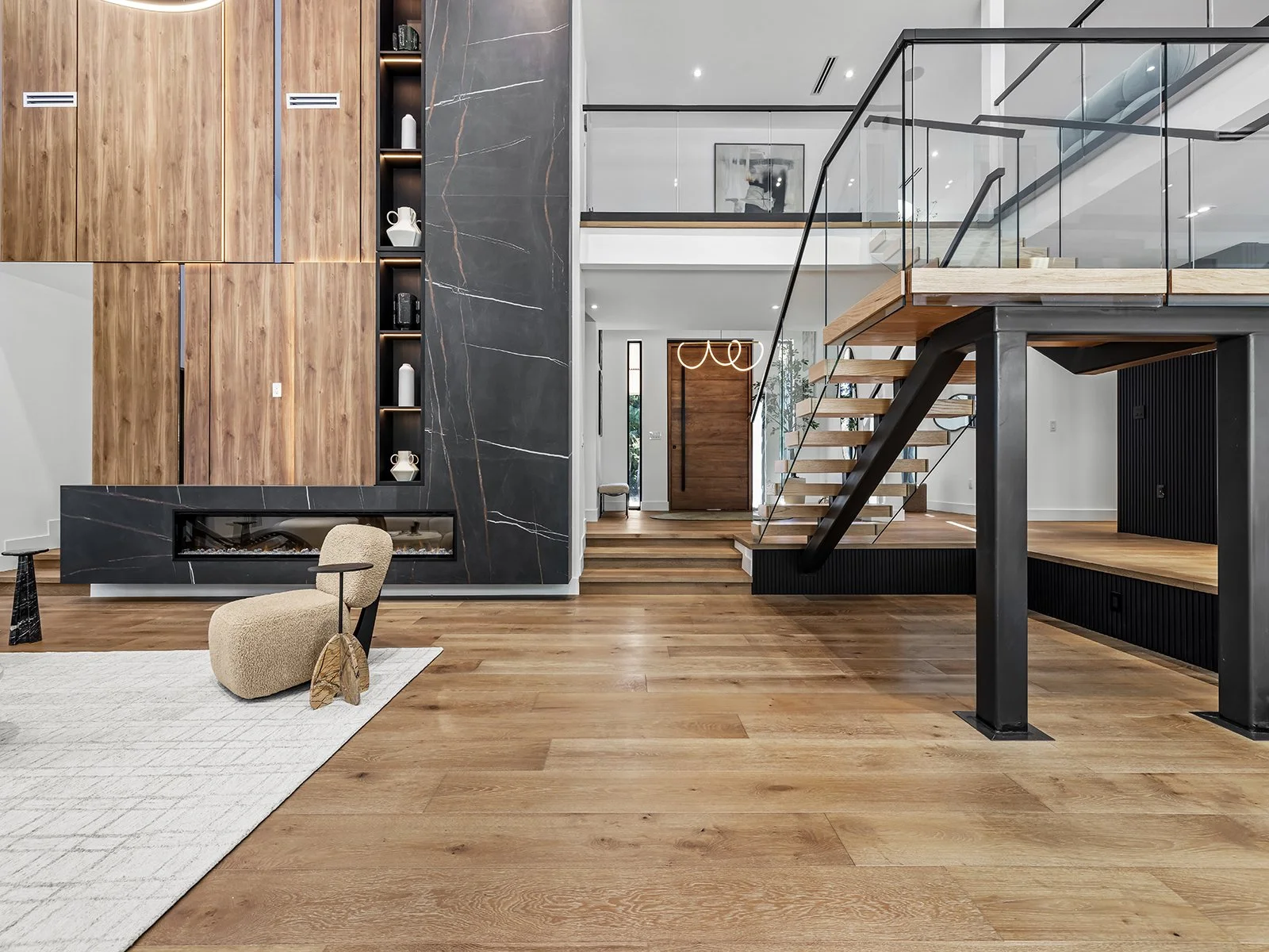 Modern living room with wooden floors, a black marble fireplace wall, and built-in wooden shelves. There is a beige lounge chair and a white rug. The space features a staircase with wooden steps and a black metal frame leading to a second level.