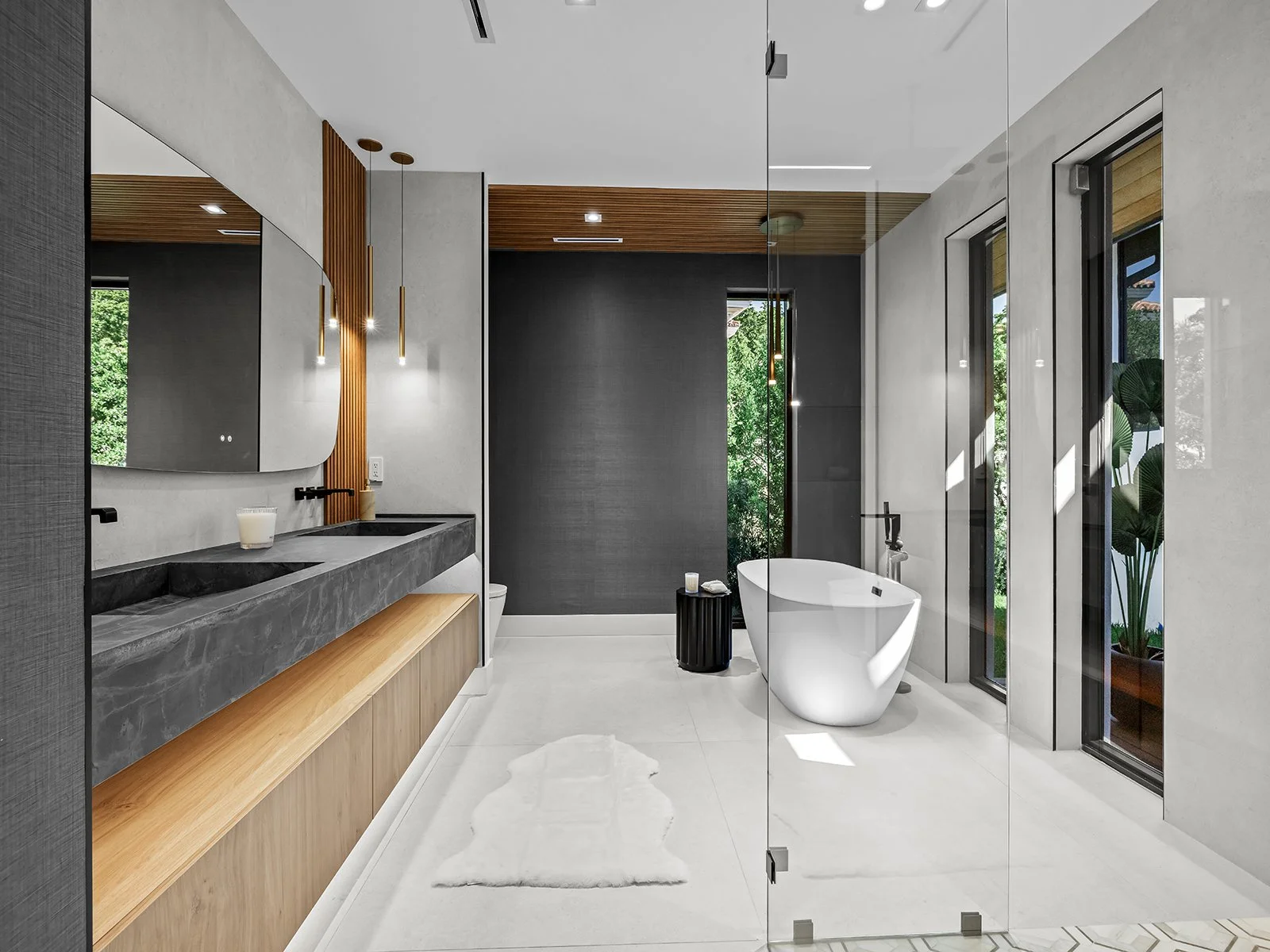 Modern bathroom with a freestanding bathtub, black and grey vanity with a mirror, and large windows with greenery outside.
