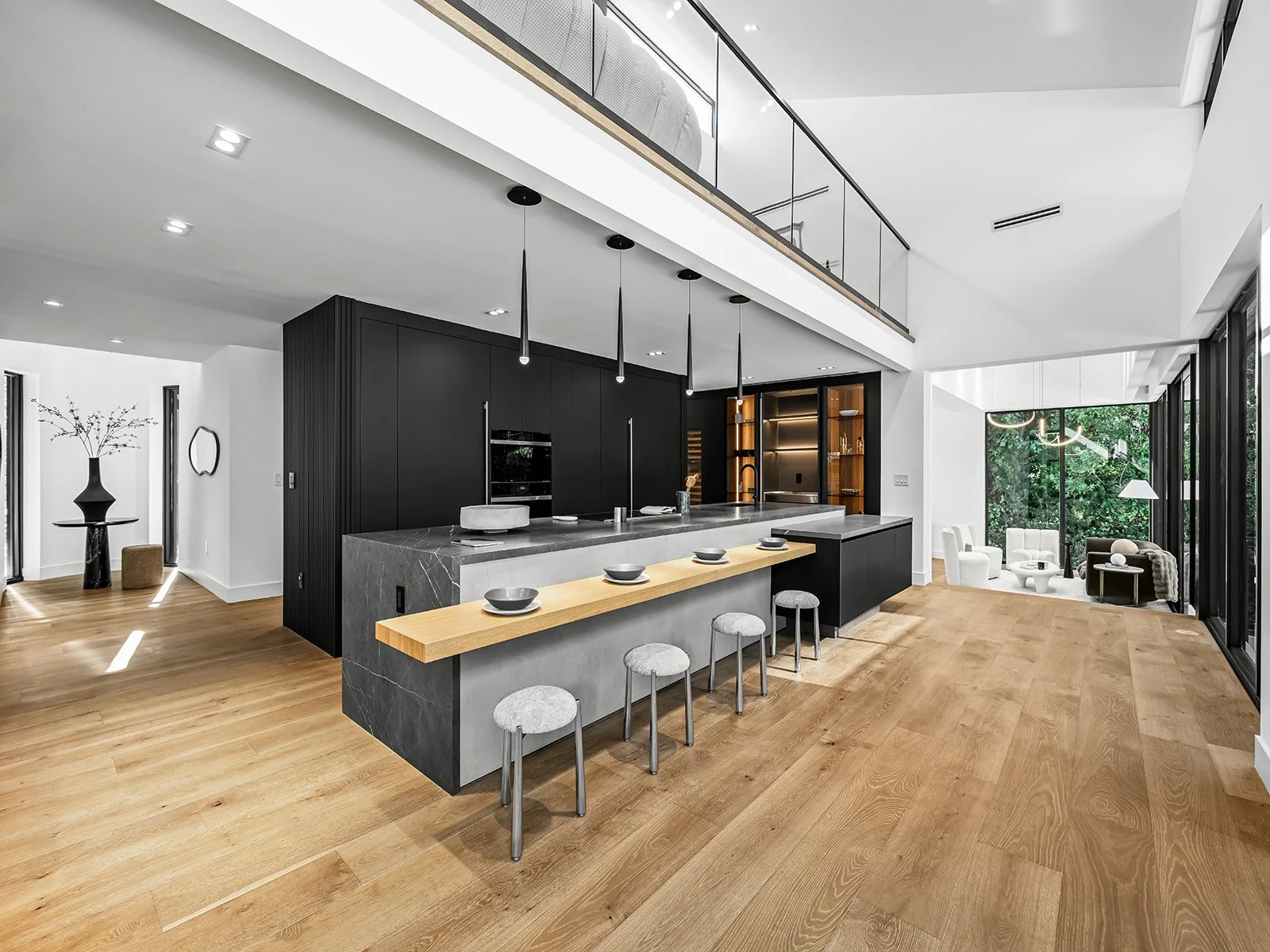 Modern open-plan kitchen with black cabinetry, gray marble island, wooden countertop, and barstools, adjacent to a living area with white and dark furniture, large windows with greenery outside, and hardwood floors.
