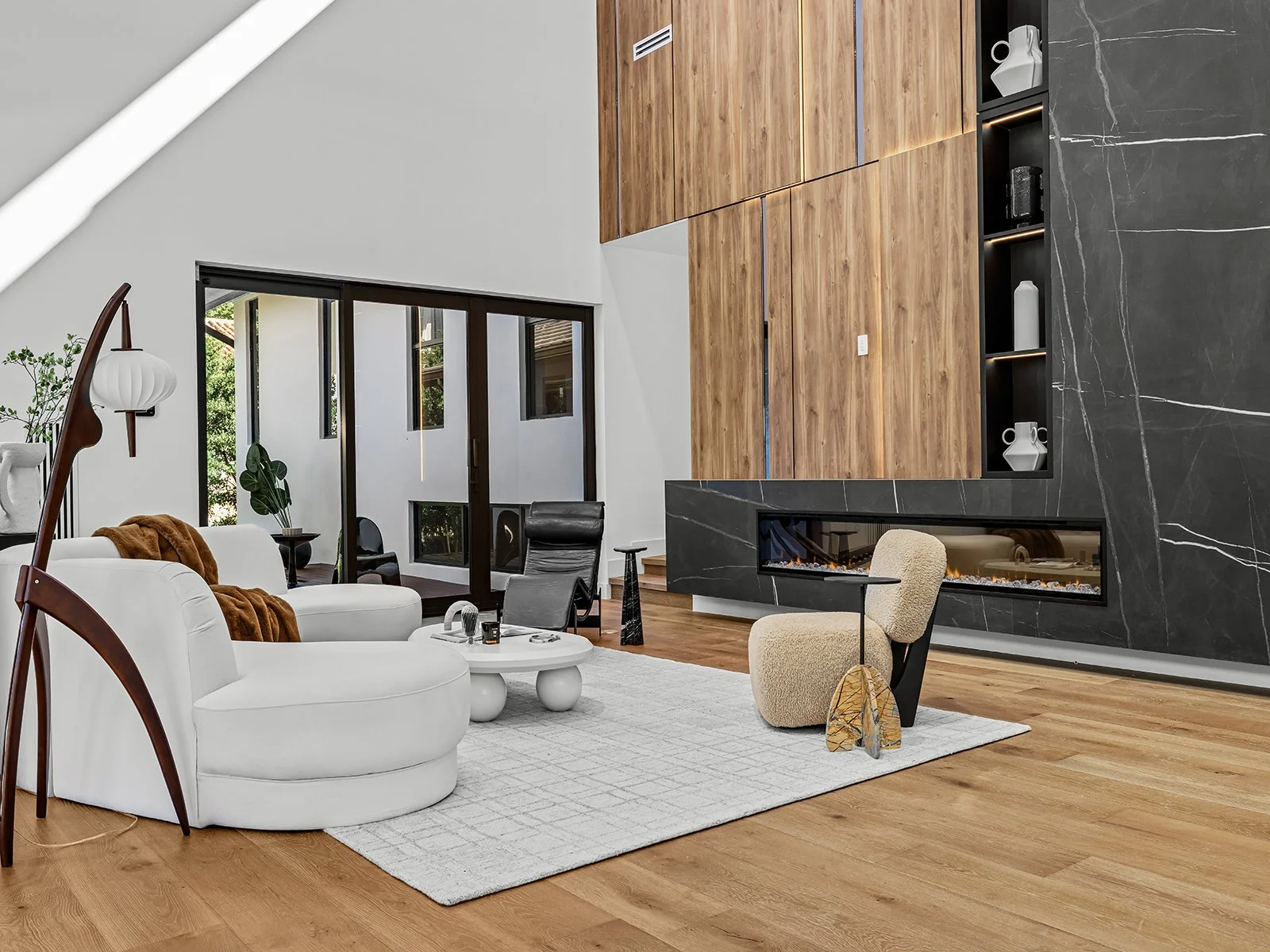 Modern living room with white sofa, black and beige chairs, fireplace, wooden and black marble wall, and large glass sliding doors leading outside.
