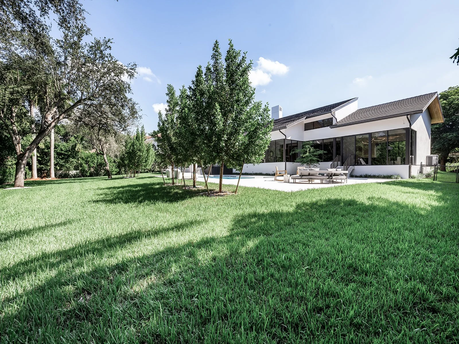 Modern white house with large glass windows, surrounded by a green lawn and trees, under a clear blue sky.