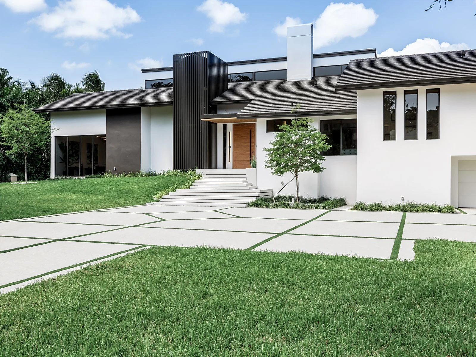 Modern two-story house with white and dark gray exterior, large windows, a small set of stairs leading to the entrance, a young tree in the front yard, and a patterned concrete driveway, under a partly cloudy sky.