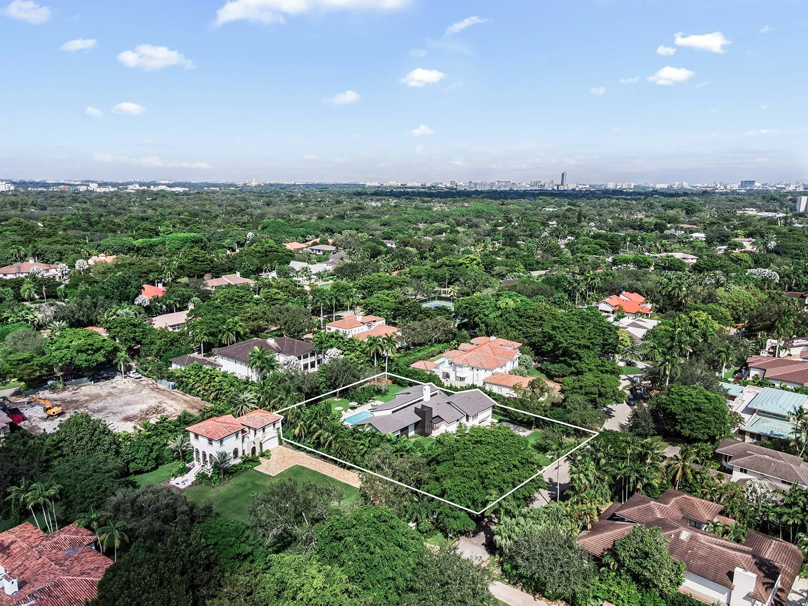 Aerial view of a suburban neighborhood with a large house outlined in white, surrounded by green trees and other residential houses, under a partly cloudy sky.
