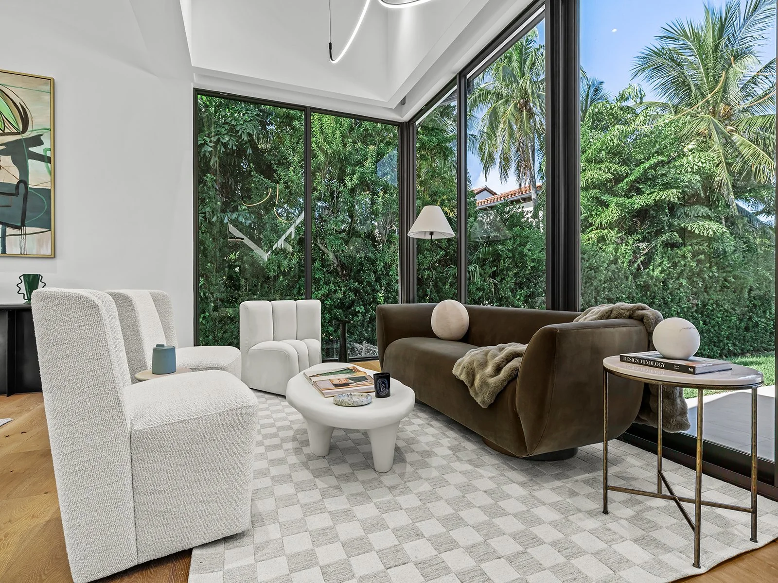 Living room with large floor-to-ceiling windows showing lush green trees outside, beige and white furniture, a brown sofa, a white textured armchair, a small round side table, and a checkered area rug.