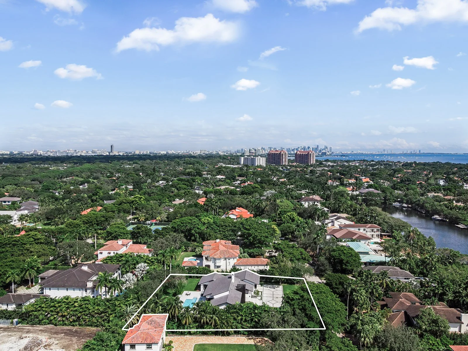 Aerial view of a residential neighborhood with houses, trees, a waterway, and a city skyline in the distance under a partly cloudy sky.