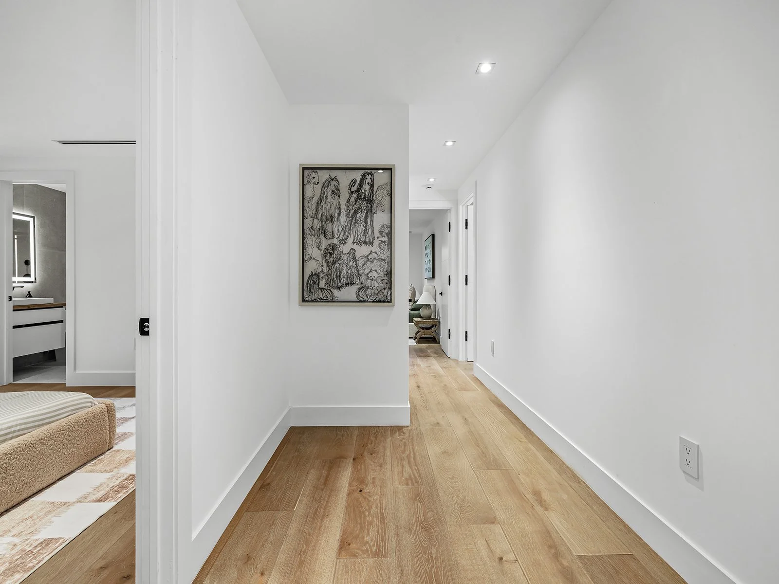White hallway with wooden floor, a framed drawing of figures on the wall, and doorways leading to other rooms.
