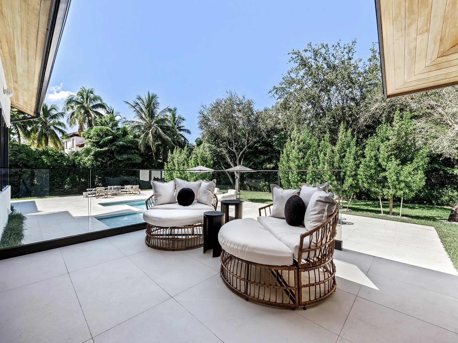 Outdoor patio with two round rattan chairs with white cushions and black pillows, small side tables, and a glass barrier overlooking a lush green yard with palm trees and a pool.